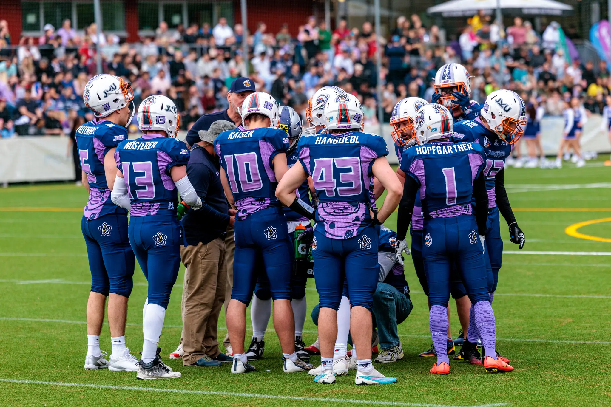 Eine Gruppe American-Football-Spieler der JKU Astros in ihren Teamuniformen bespricht die Strategie auf dem grünen Rasenfeld 