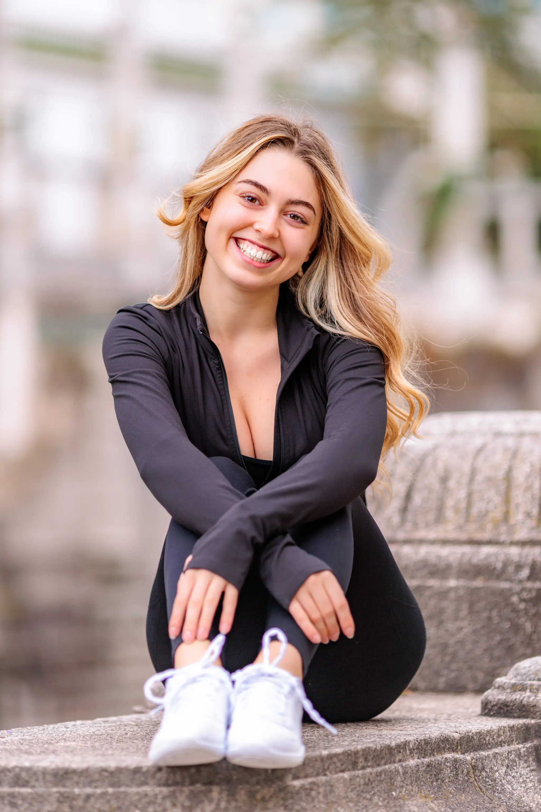 Model Valentina sitzt in einem sportlichen schwarzen Outfit auf einer Steinstufe und lacht während ihres Fotoshootings im Stadtpark herzlich in die Kamera