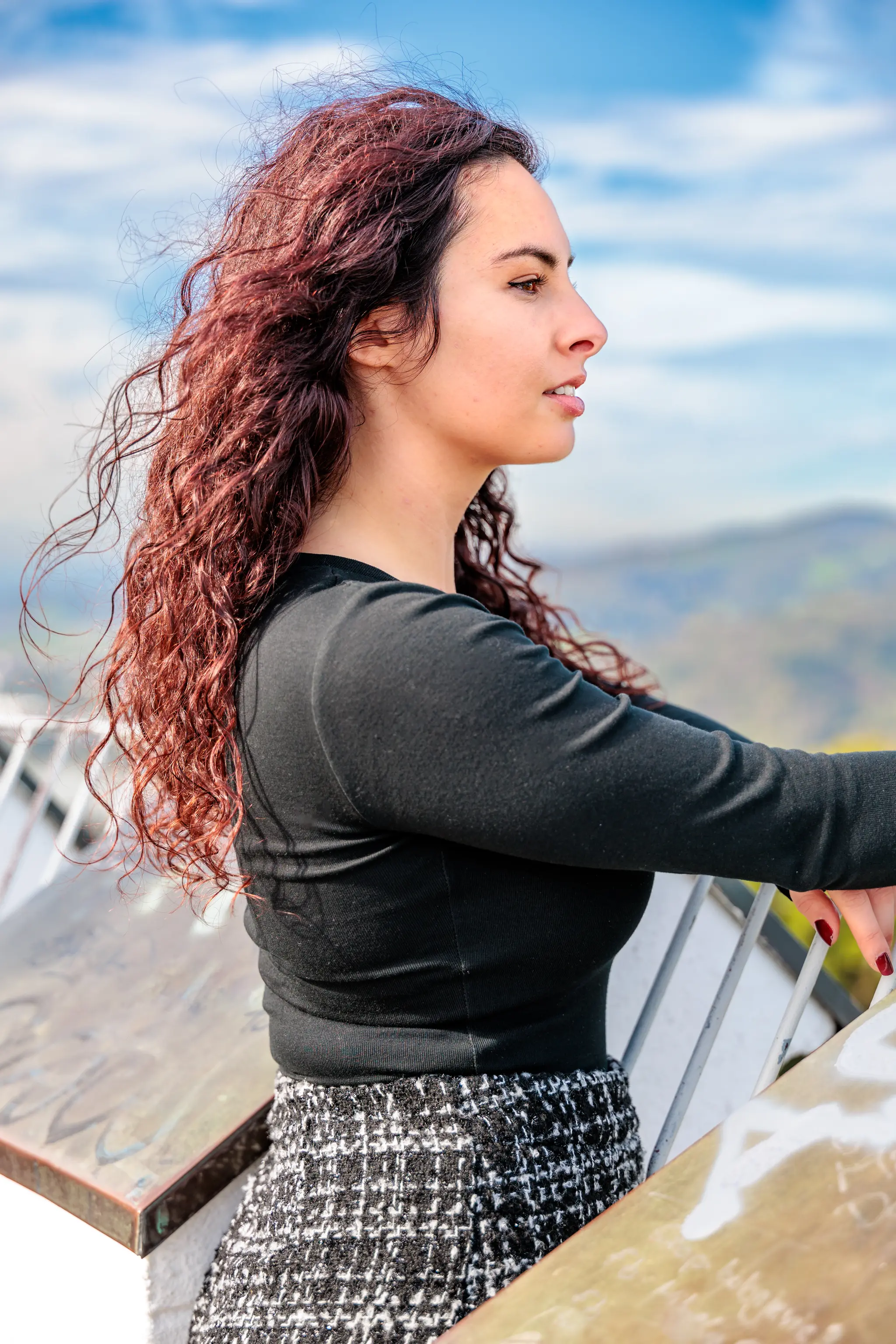 Eine junge Frau mit rotbraunem Lockenhaar blickt im Profil von einem Aussichtspunkt auf eine weite Landschaft unter blauem Himmel 