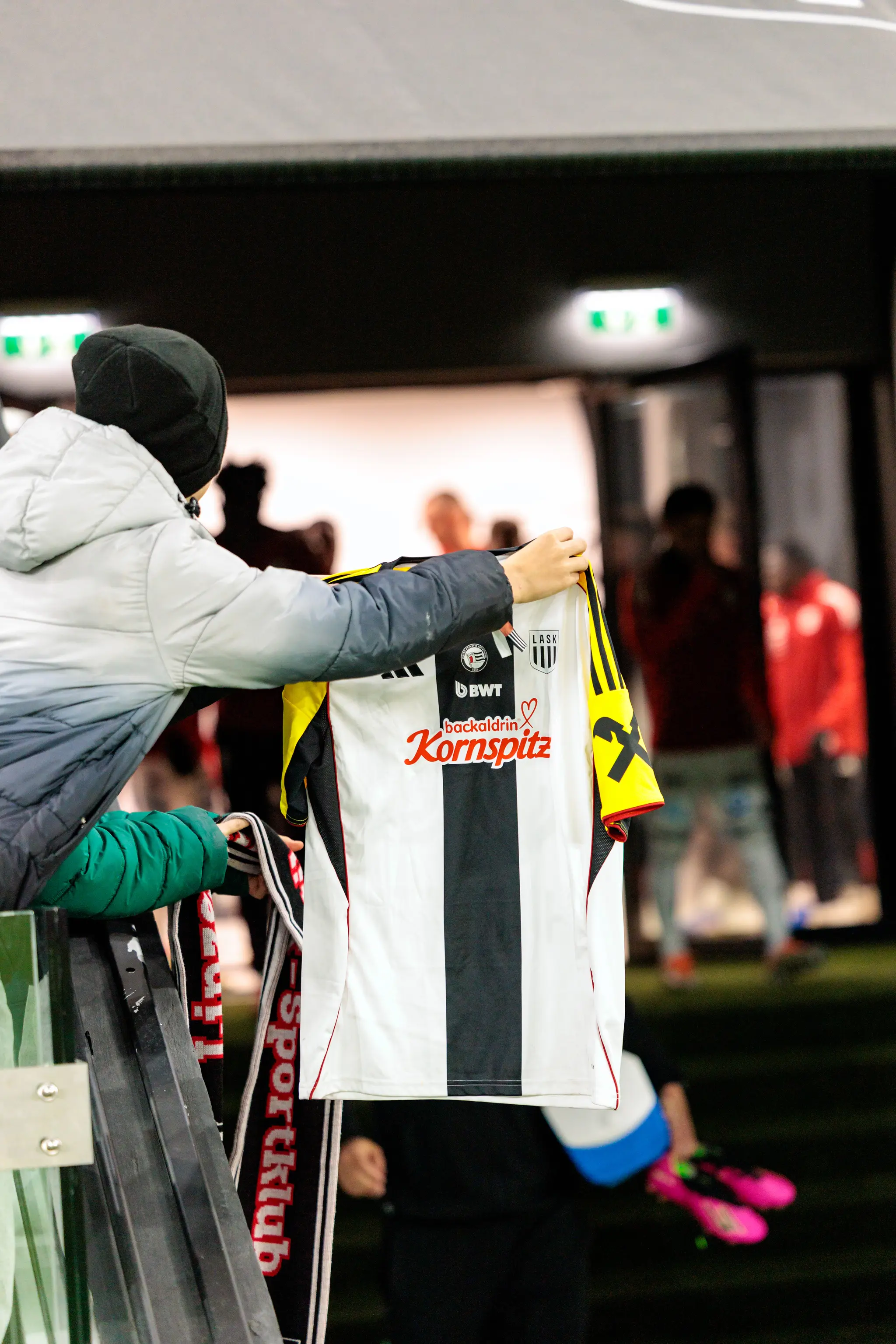 Ein LASK-Fan in grauer Jacke und schwarzer Mütze hält ein weiß-schwarz gestreiftes Fußballtrikot des LASK mit den Sponsoren BWT und Kornspitz an einem Geländer im Stadion hoch