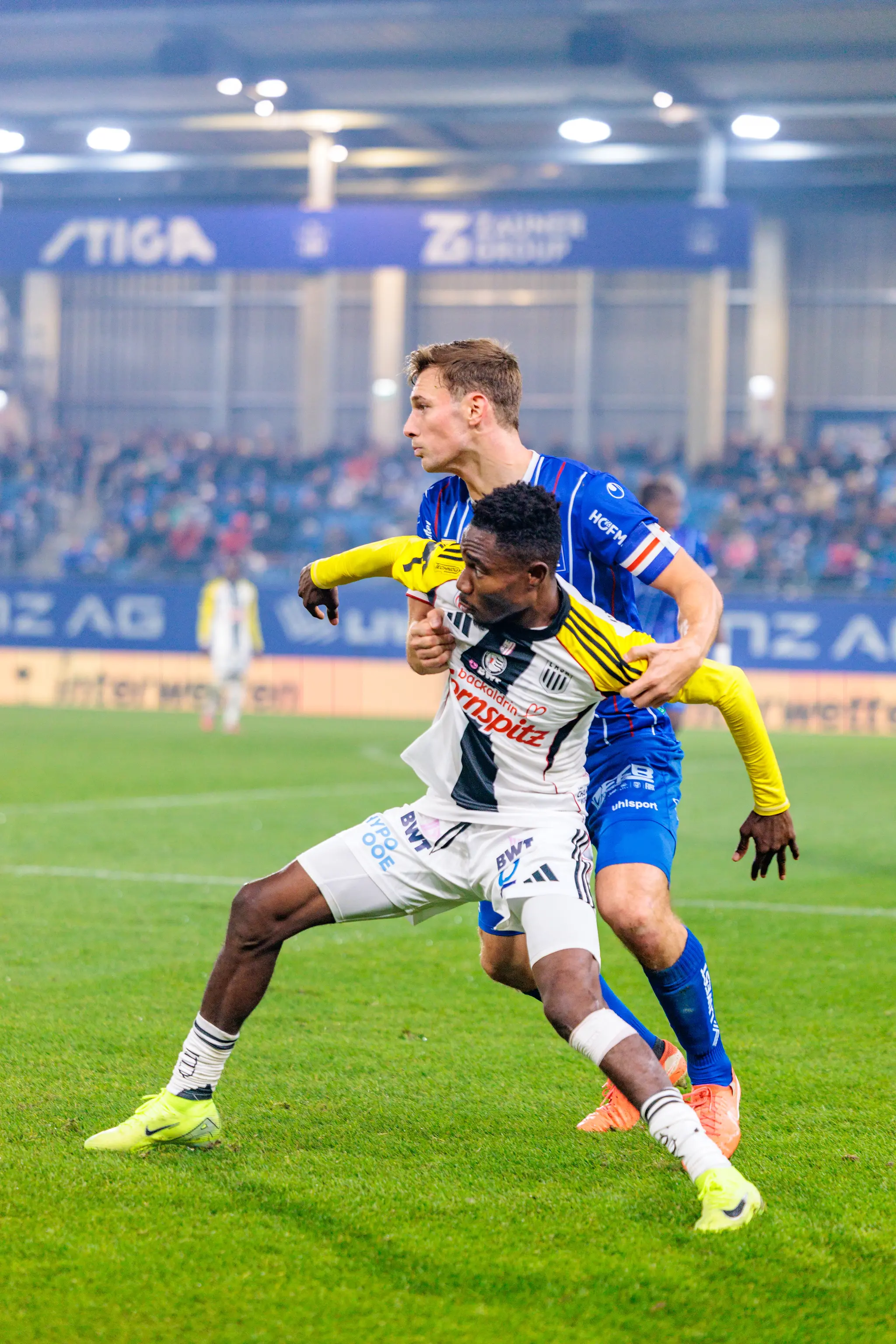 LASK-Spieler George Bello im schwarz-weiß gestreiften Trikot wird von einem Spieler von Blau-Weiß Linz im blauen Trikot in einem intensiven Zweikampf festgehalten