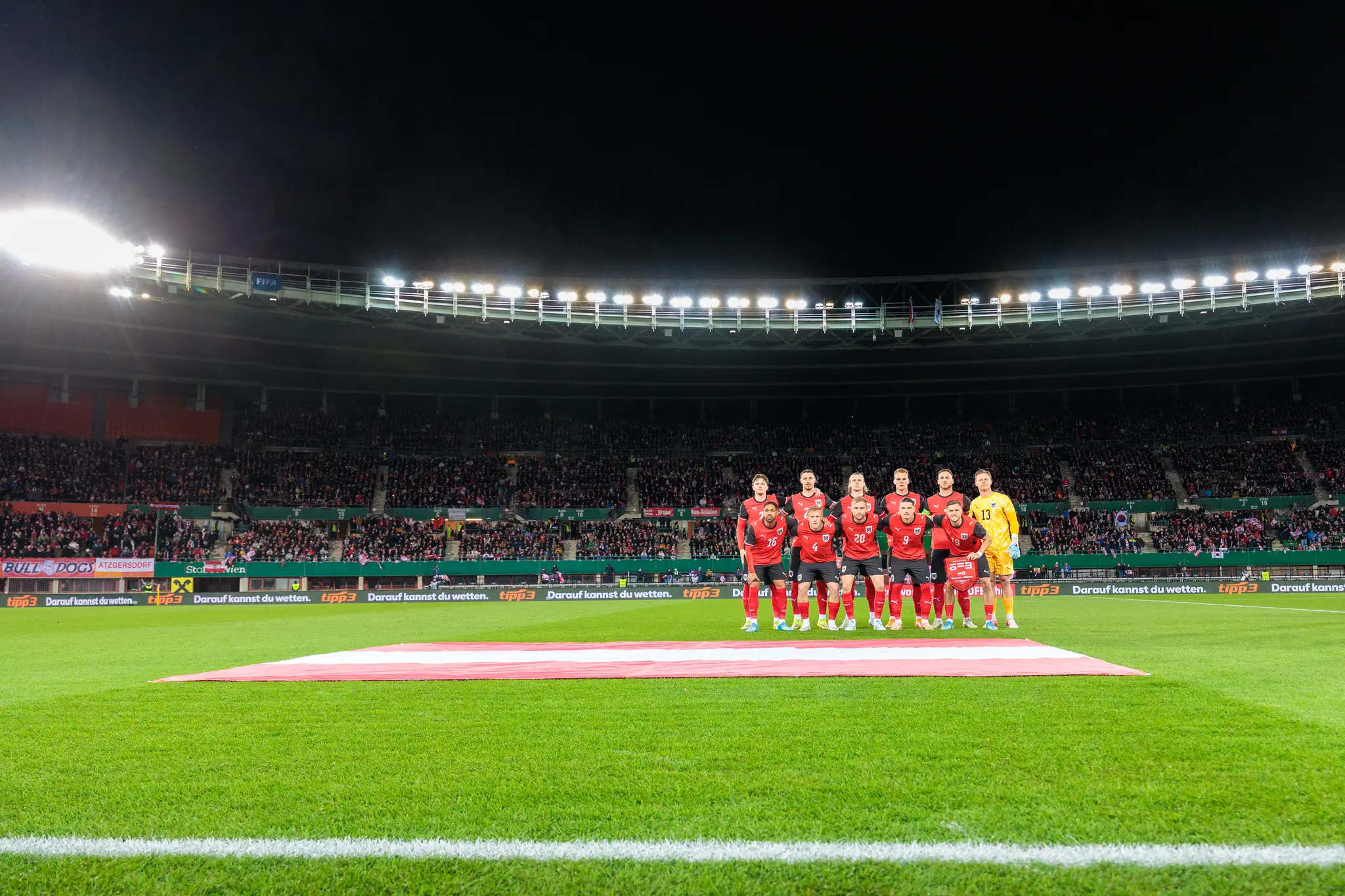 Das österreichische Fußballnationalteam posiert in roten Trikots vor der österreichischen Flagge und gefüllten Zuschauerrängen im hell erleuchteten Happel Stadion