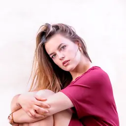 Eine junge Frau mit langen braunen Haaren in einem dunkelroten Oberteil sitzt mit um die Knie geschlungenen Armen und blickt nachdenklich in die Kameralens: 56mm F1.4 DC DN | Contemporary 018