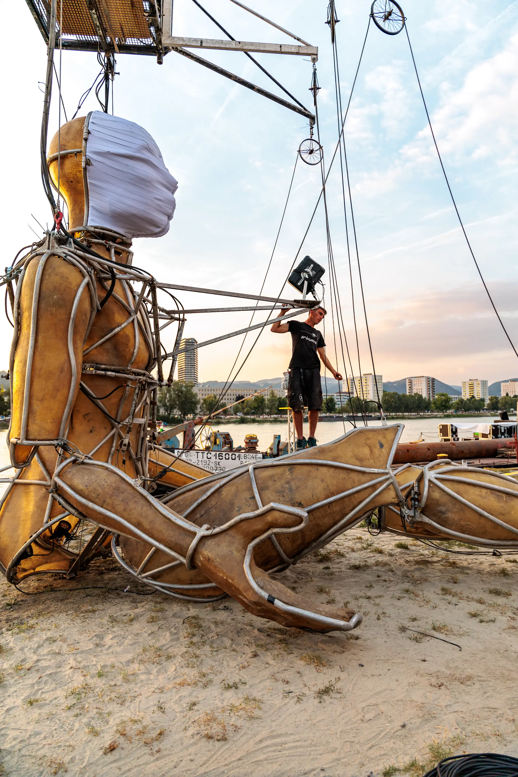 Riesige goldene Gliederpuppe mit weißer Gesichtsmaske an einem Flussufer, daneben ein Techniker, der eine Seilkonstruktion und Beleuchtungselemente justiert, im warmen Licht des Sonnenuntergangs mit Stadtsilhouette im Hintergrund 