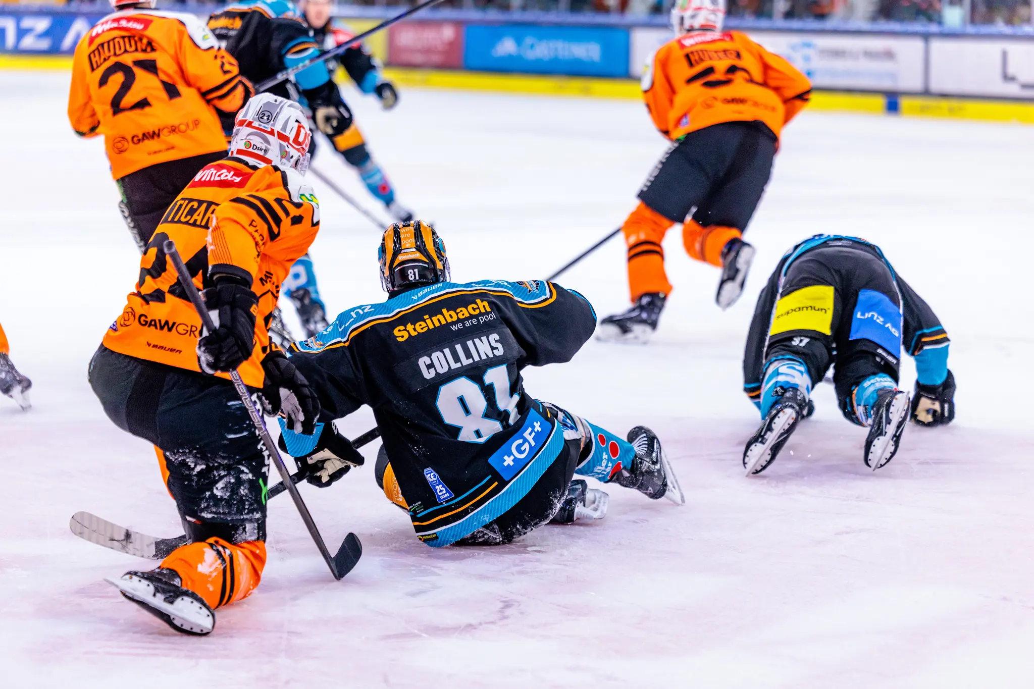 Eishockey-Spieler in orangefarbenen und schwarz-türkisen Trikots kämpfen auf dem Eis, wobei zwei Spieler der Black Wings Linz, darunter der mit der Nummer 81, am Boden liegen 