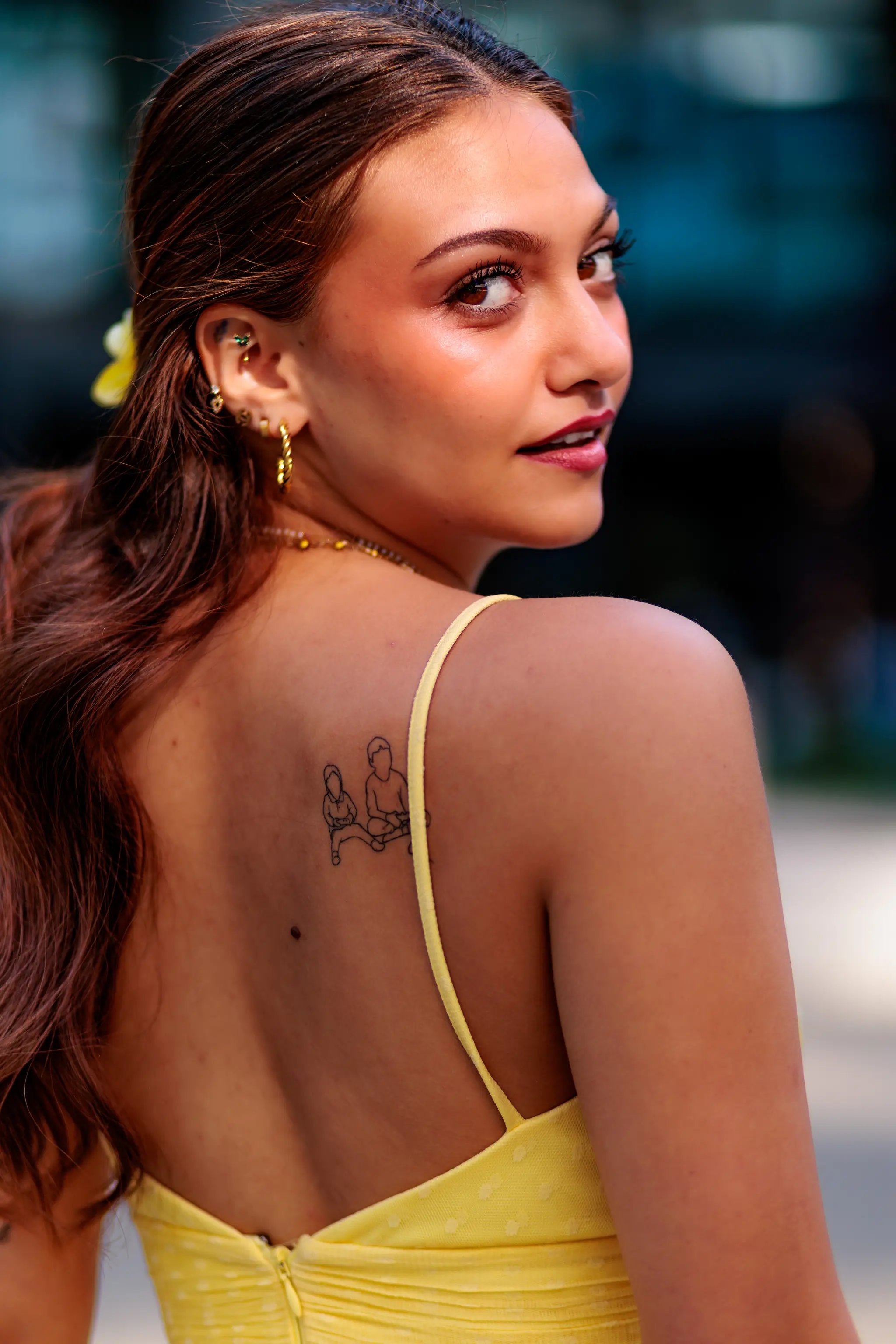 Nahaufnahme einer jungen Frau mit braunem Haar, das zu einer Seite getragen wird, und einem Tattoo auf ihrem oberen Rücken. Sie trägt ein gelbes Kleid und hat mehrere Ohrringe. 