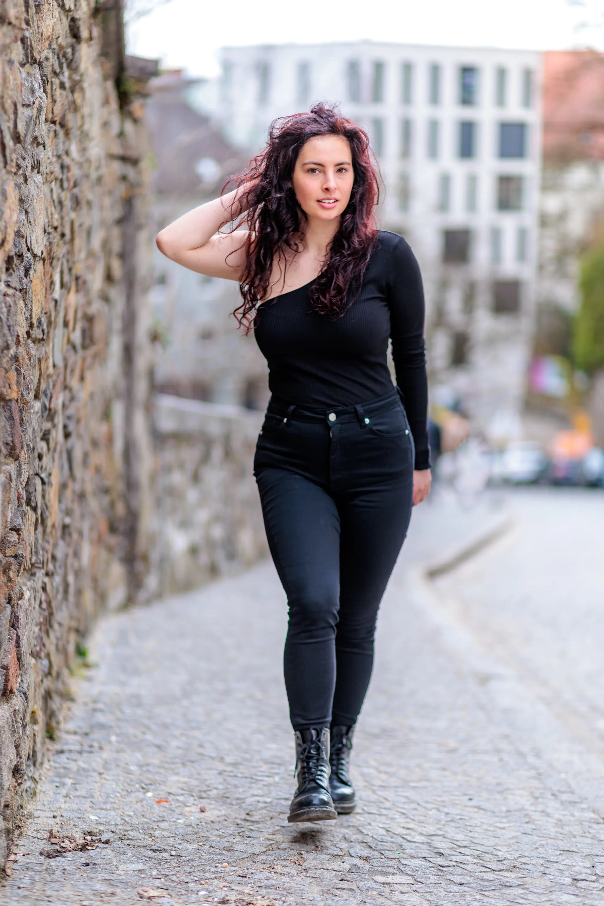 Porträt einer jungen Frau mit lockigem Haar in schwarzer Kleidung, selbstbewusst auf gepflasterter Gasse mit Steinmauer und urbanem Hintergrund