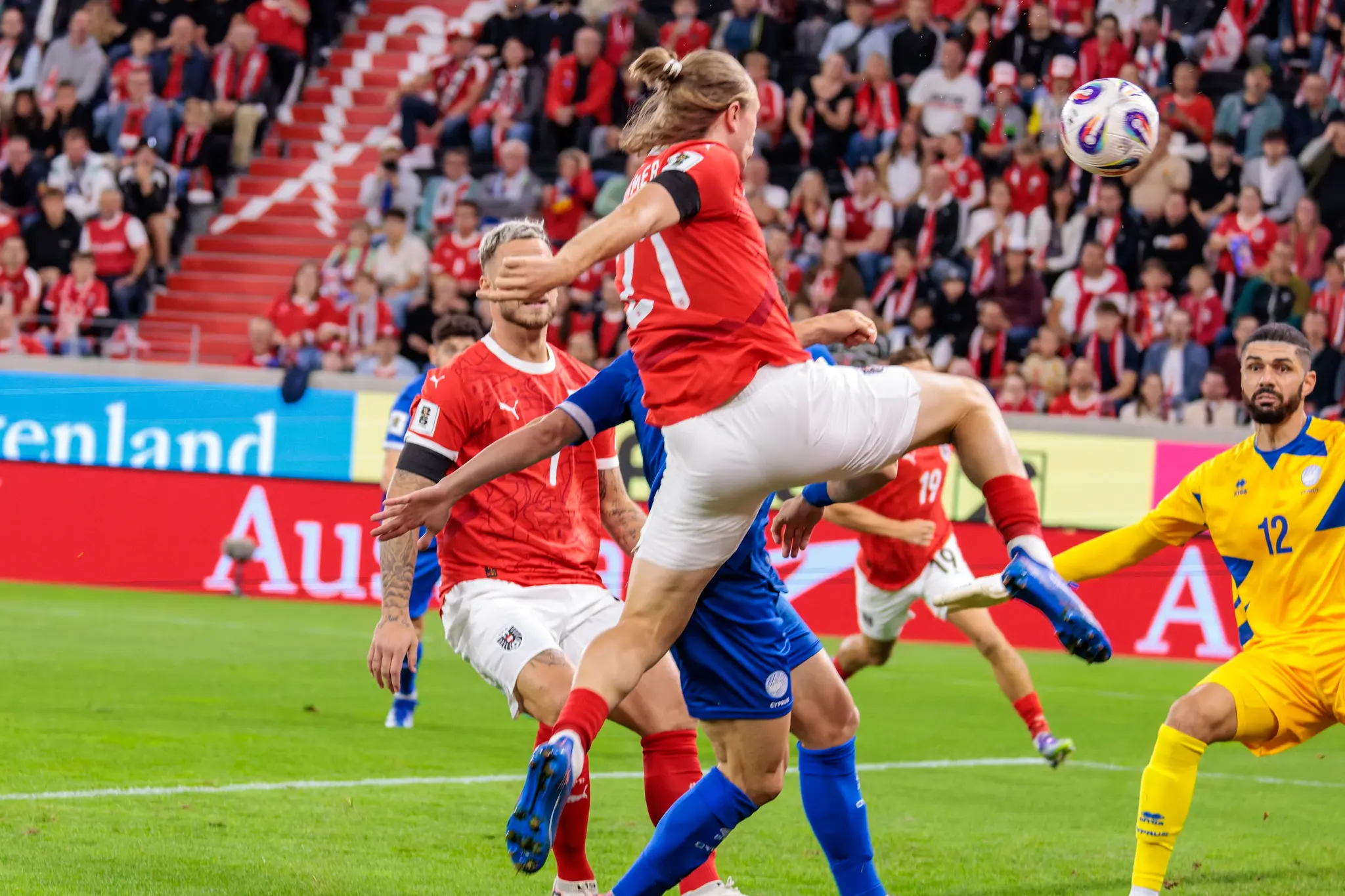 Ein österreichischer Spieler mit der Rückennummer 21, Patrick Wimmer, steigt im Strafraum zum Ball hoch, während der zypriotische Torhüter Fabiano mit der Nummer 12 das Spiel aufmerksam verfolgt