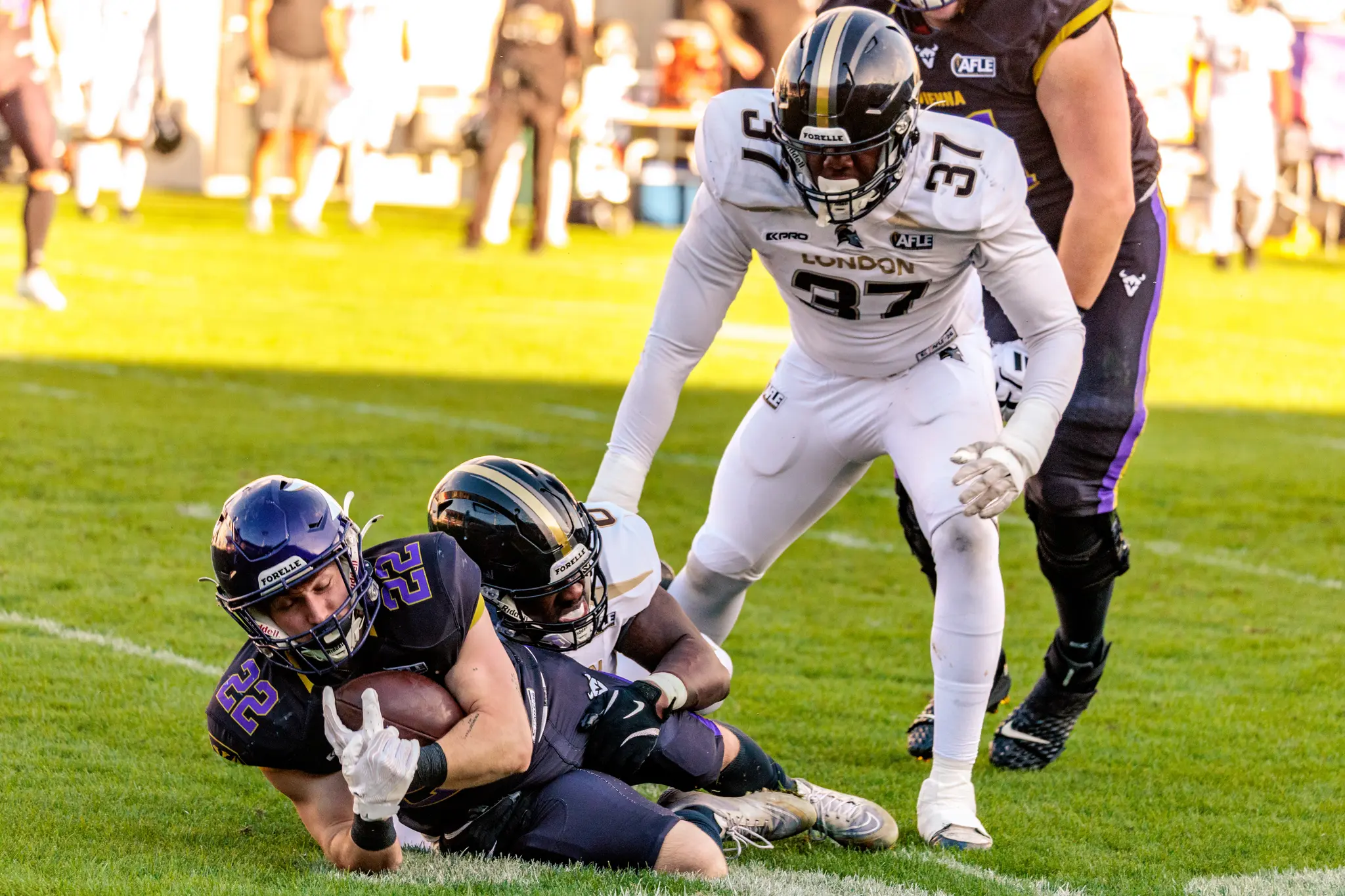 Albert Wiesigstrauch (22) liegt nach einem Tackle am Boden und sichert den Ball, darüber steht London-Spieler Nr. 37 – hartes Laufspiel der Vienna Vikings im ersten Viertel.