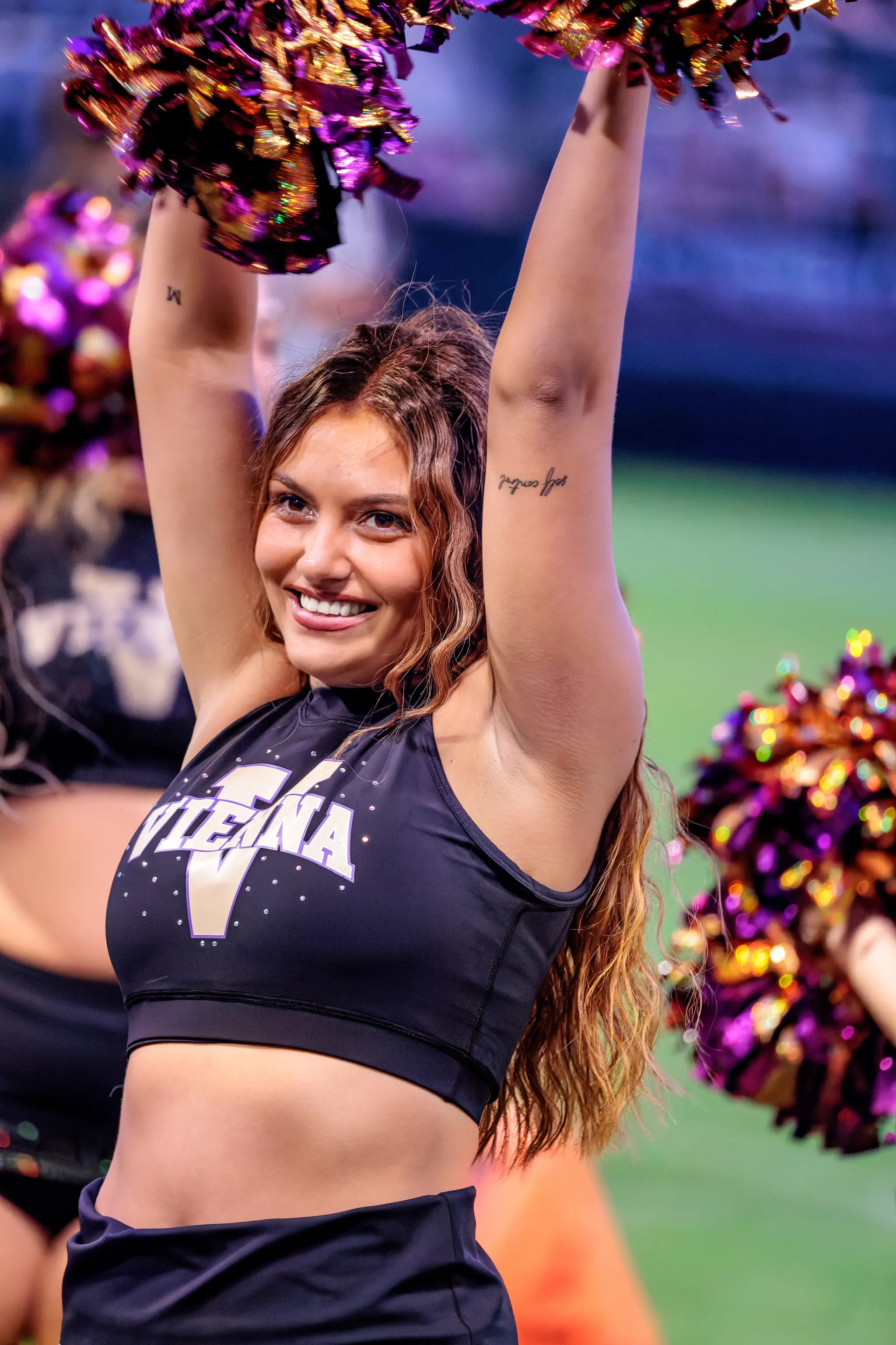 Cheerleaderin des Spirit Squad jubelt mit beiden Armen über dem Kopf und violett-goldenen Pompons.