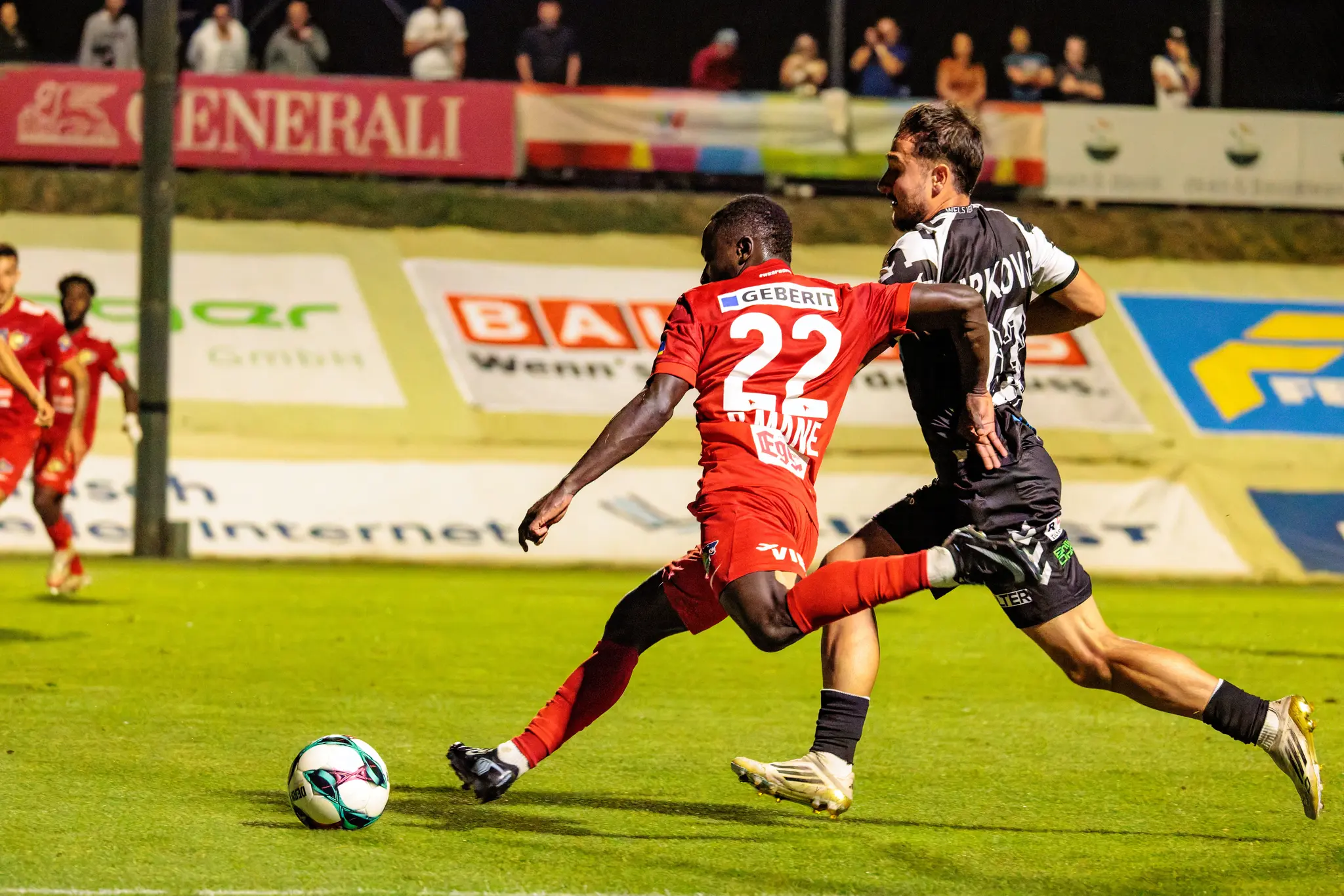 Ein dynamischer Zweikampf am Spielfeldrand zwischen einem Spieler von Herta Wels im roten Trikot mit der Nummer 22 und einem Spieler des SKN St. Pölten im schwarz-weißen Trikot