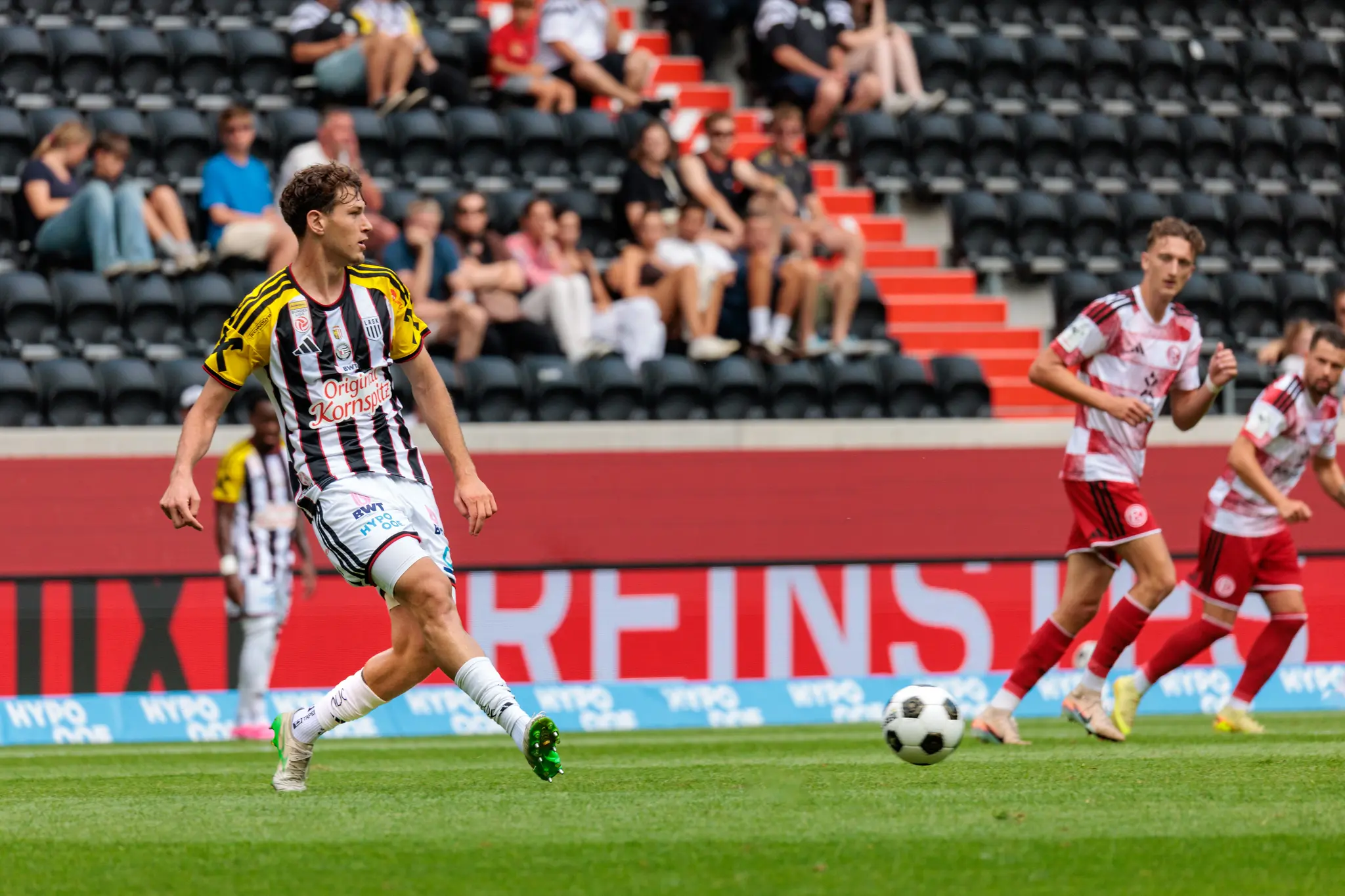 LASK-Spieler im schwarz-weiß-gelben Streifentrikot im Laufduell mit dem Ball, im Hintergrund zwei Düsseldorfer Spieler und Zuschauer auf den Rängen.
