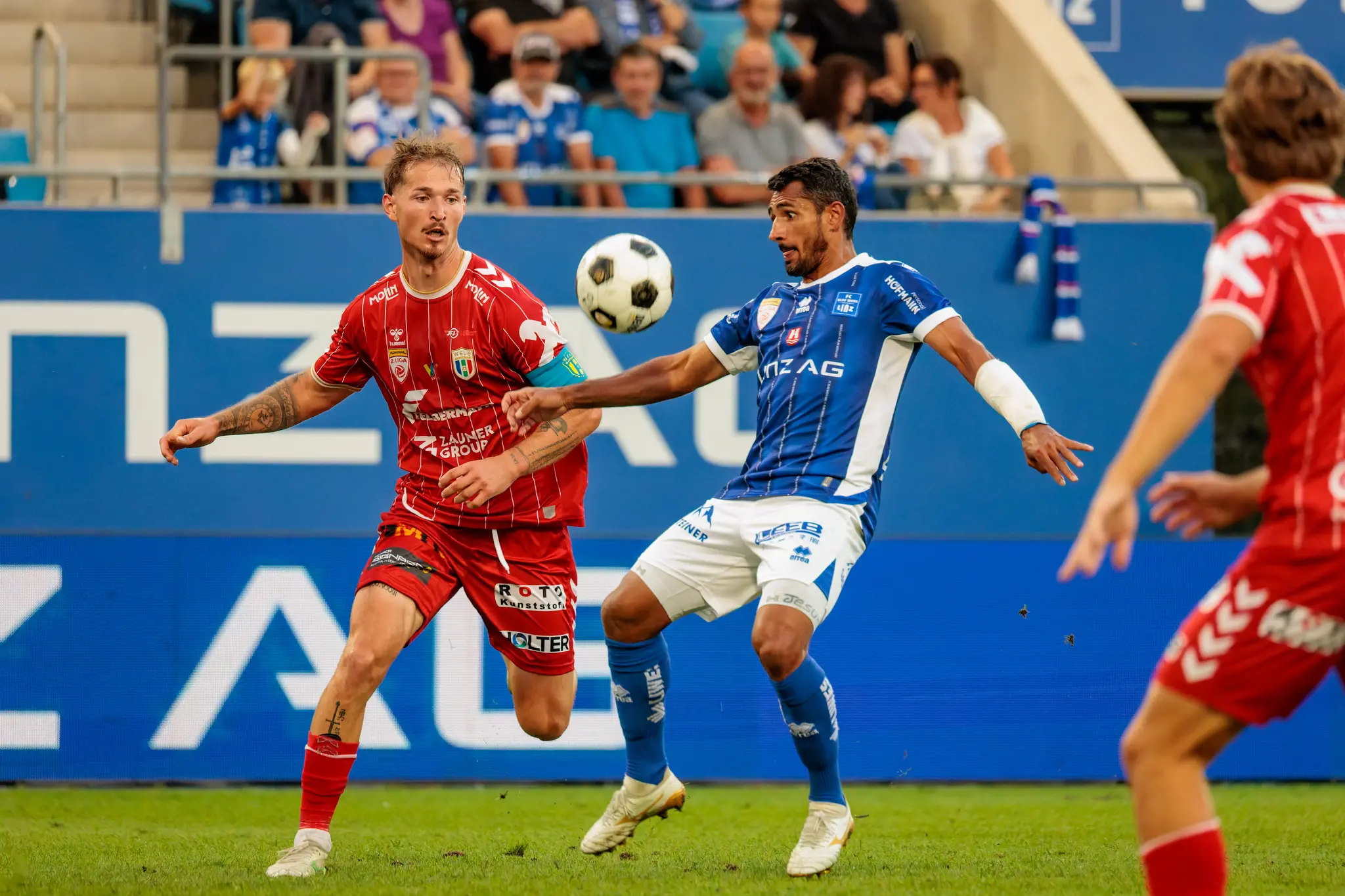 Ronivaldo vom FC Blau-Weiß Linz und ein Welser Spieler mit Kapitänsbinde ringen in der Luft um den Ball, der auf Brusthöhe schwebt; im Hintergrund ist die Linz-AG-Bande zu sehen.