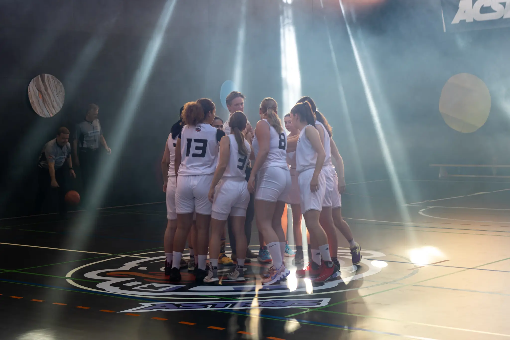 Ein Team von Basketballspielerinnen in hellen Trikots bespricht sich im Kreis auf dem Spielfeld einer Sporthalle unter atmosphärischer Beleuchtung