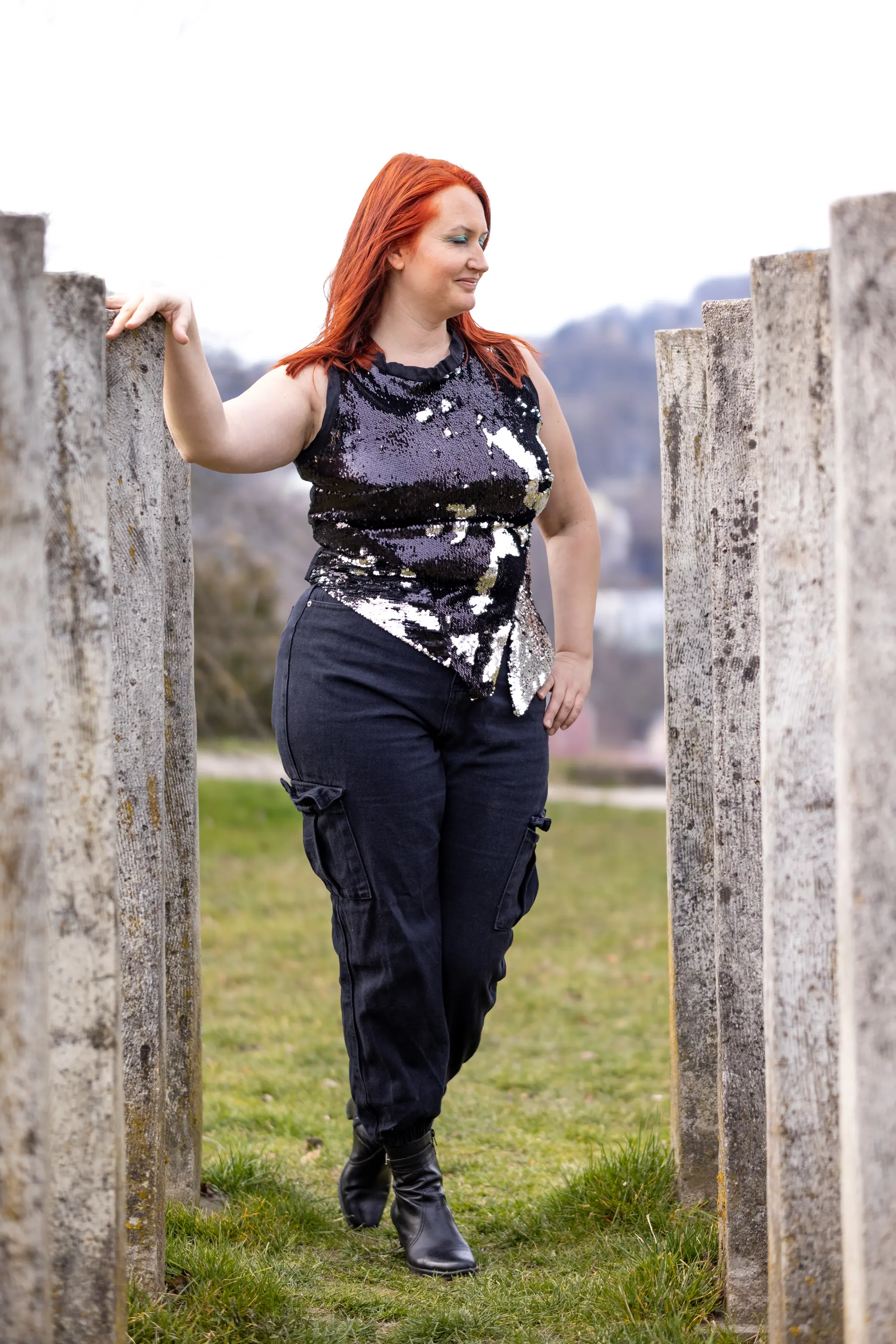 Eine rothaarige Frau in einem schwarz-silbernen Pailletten-Oberteil und dunklen Hosen lächelt, während sie sich im Freien an einem Betonpfeiler festhält