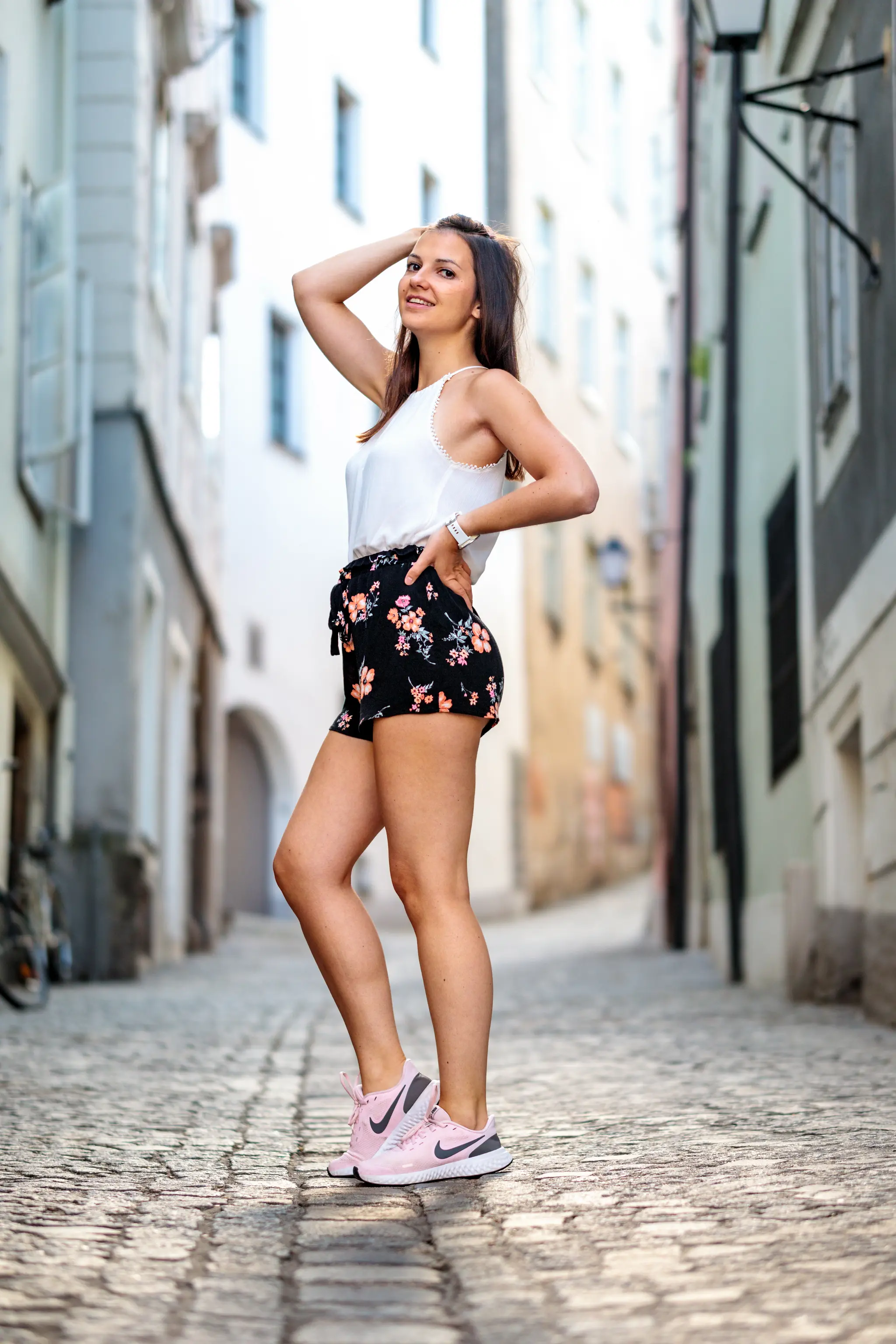 Eine lächelnde junge Frau in einer weißen Bluse, Blumenshorts und pinken Nike-Schuhen posiert in einer gepflasterten Altstadtgasse