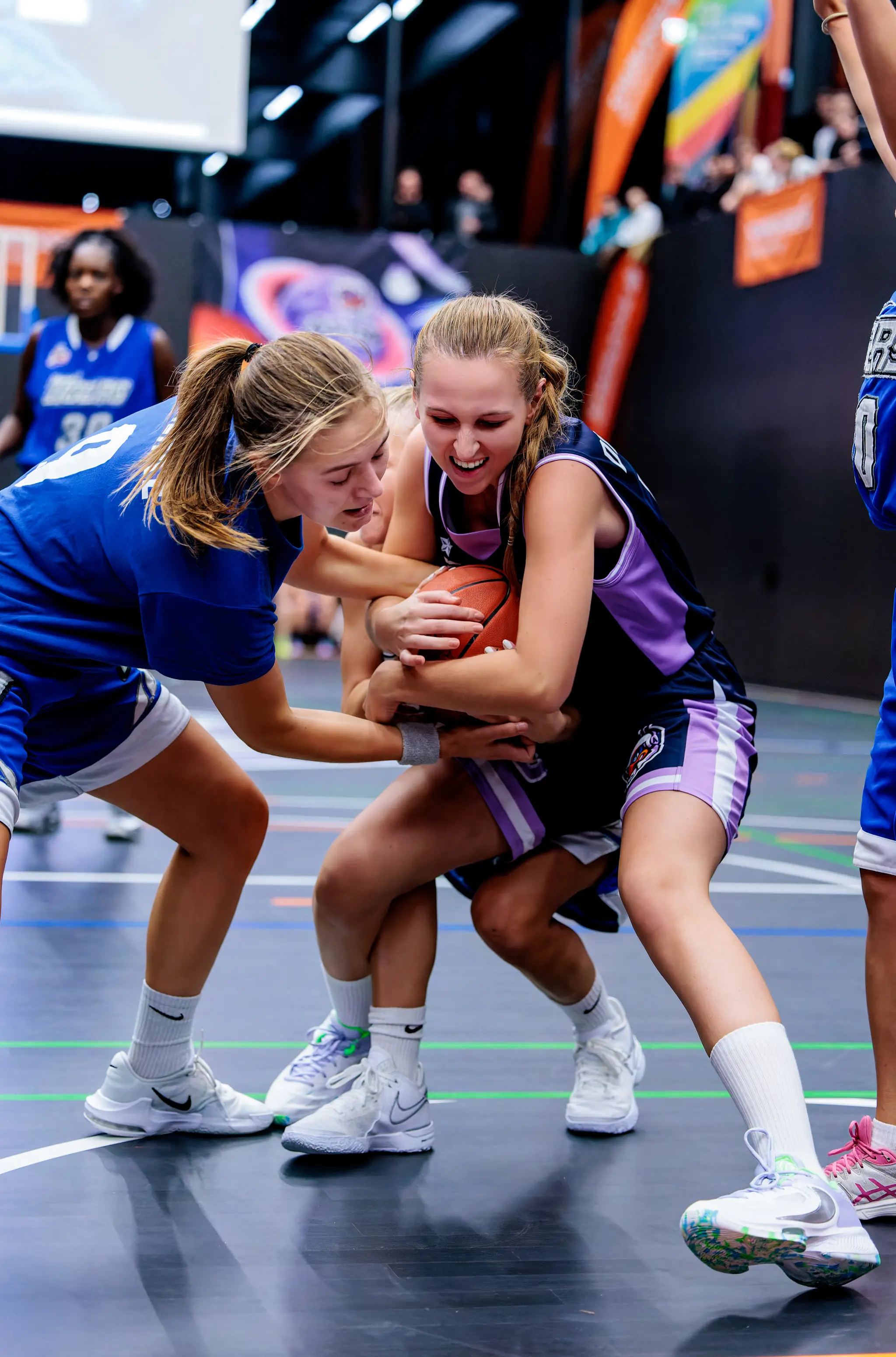 Zwei junge Basketballspielerinnen in blauen und lila Trikots kämpfen energisch um den Ball auf einem glänzenden Hallenplatz