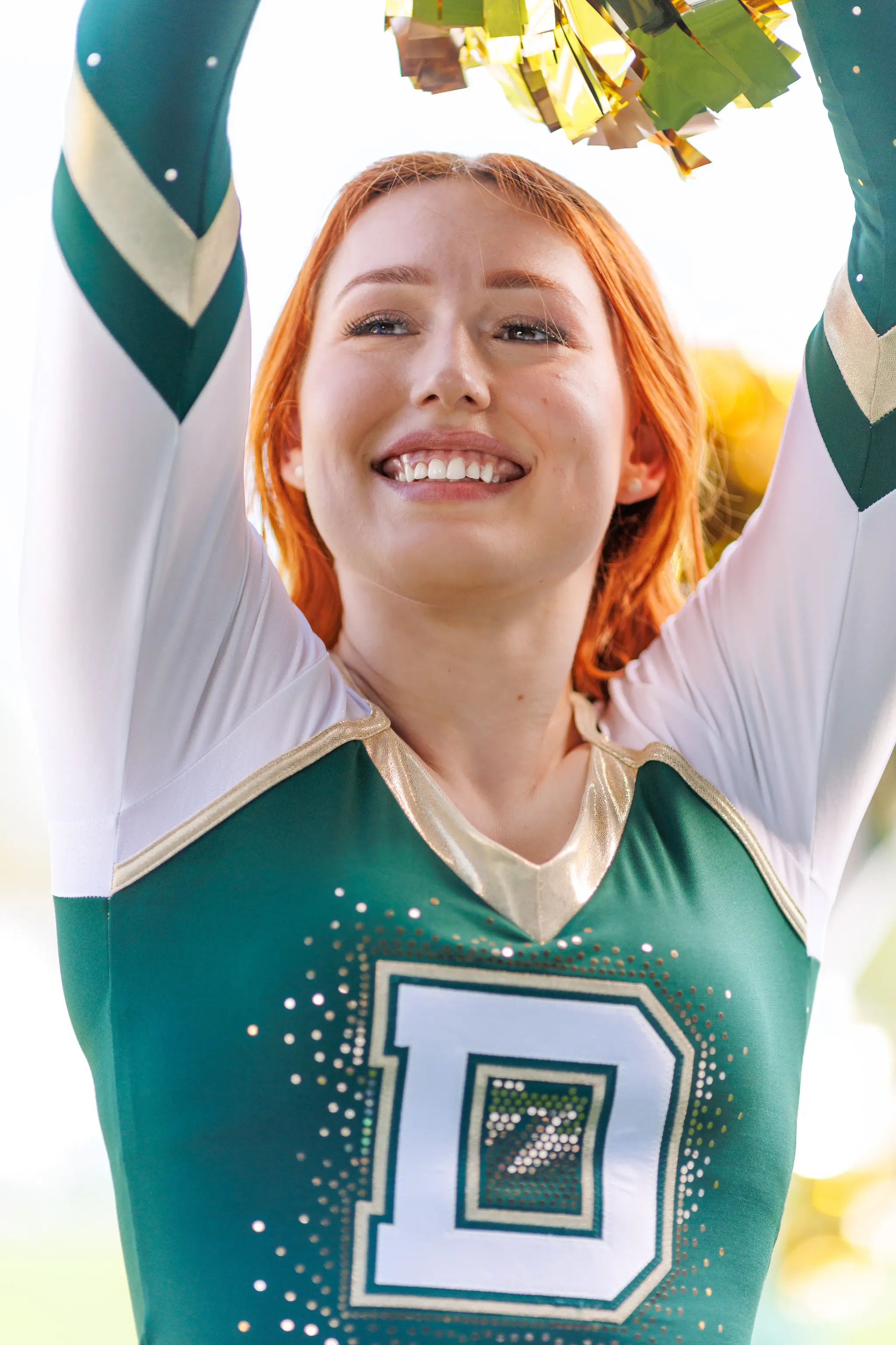 Eine lachende Cheerleaderin der Danube Dragons mit rotem Haar hält goldene Pompoms in ihrer glitzernden grünen und weißen Uniform hoch
