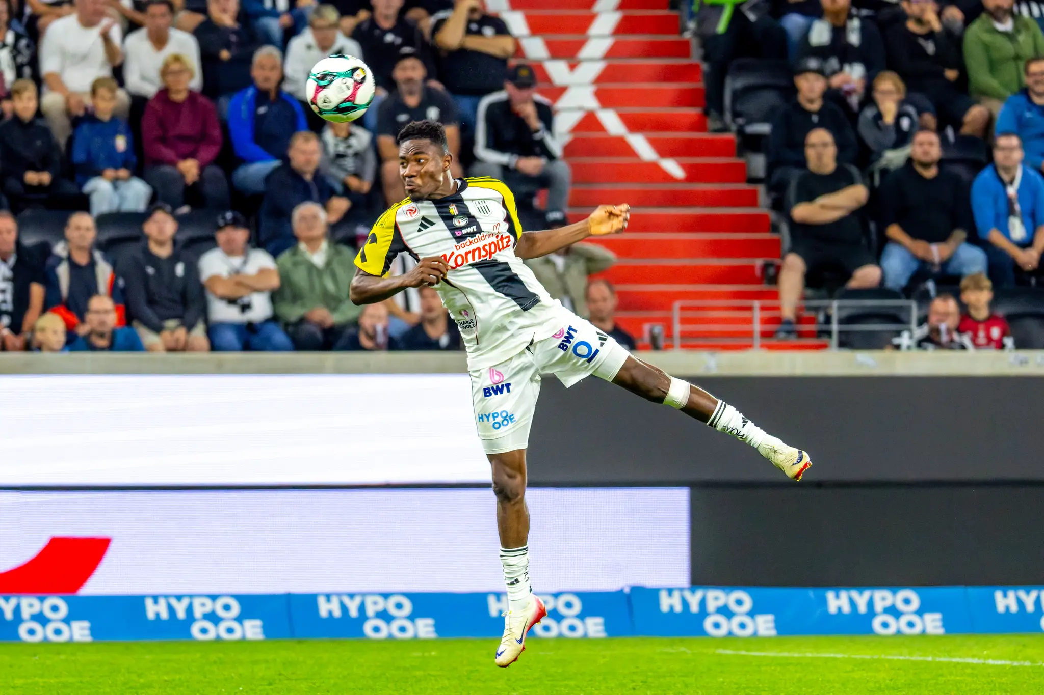 Ein LASK-Spieler im weiß-gelben Trikot springt im Stadion zum Kopfball, während Fans auf den Rängen zu sehen sind