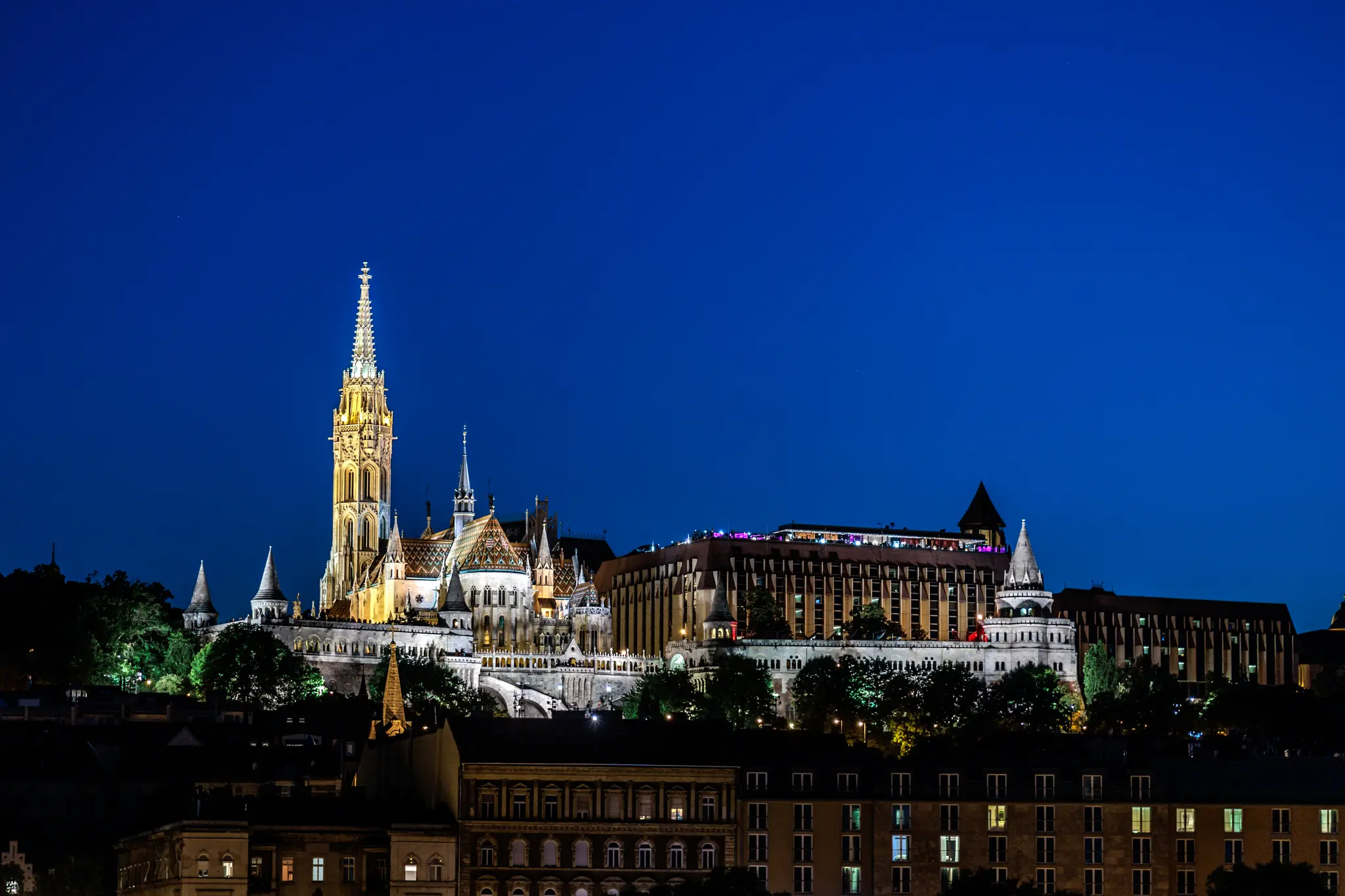 Nachtansicht von Budapest mit der hell beleuchteten Matthiaskirche und Fischerbastei auf dem Burgberg vor tiefblauem Himmel. 