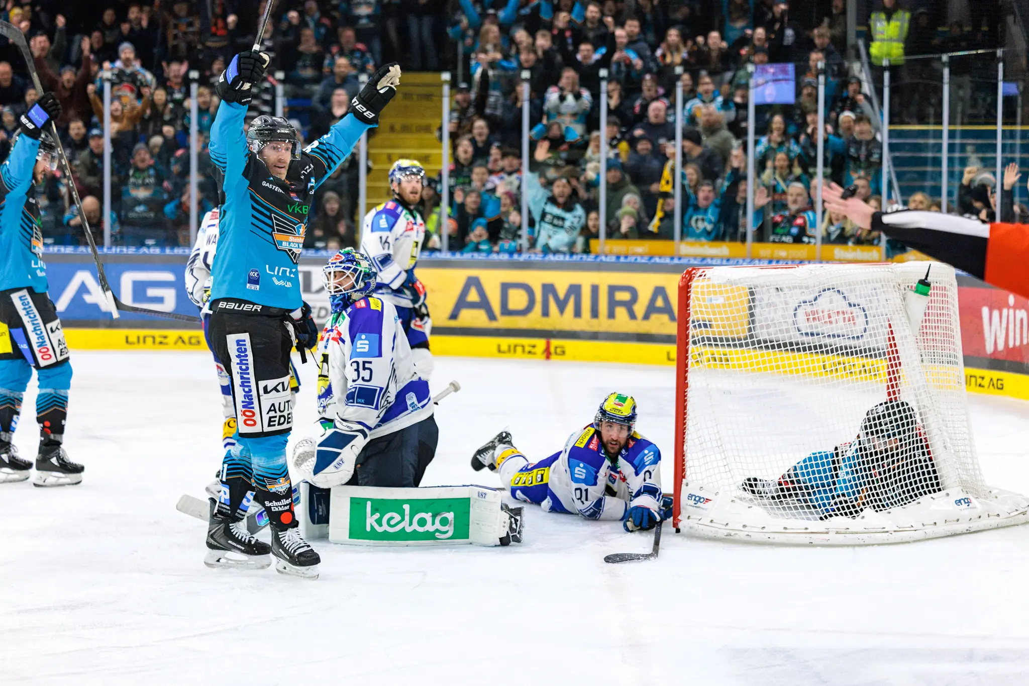 Ein jubelnder Eishockeyspieler der Black Wings Linz mit erhobenen Armen nach einem Tor, während der VSV-Torhüter enttäuscht am Boden kniet und die Fans im Hintergrund jubeln