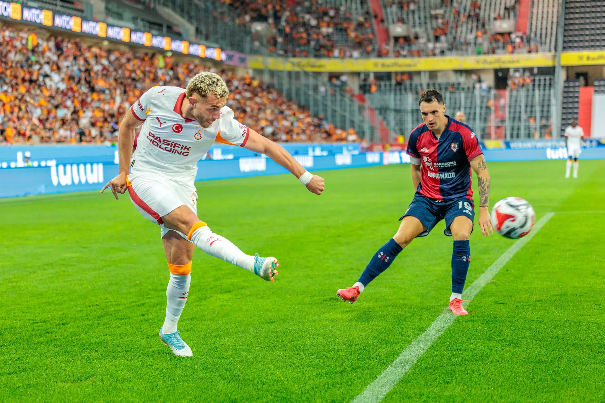 Ein Galatasaray-Fußballspieler in weißem Trikot schießt den Ball in einem Stadion, beobachtet von einem Cagliari-Spieler 