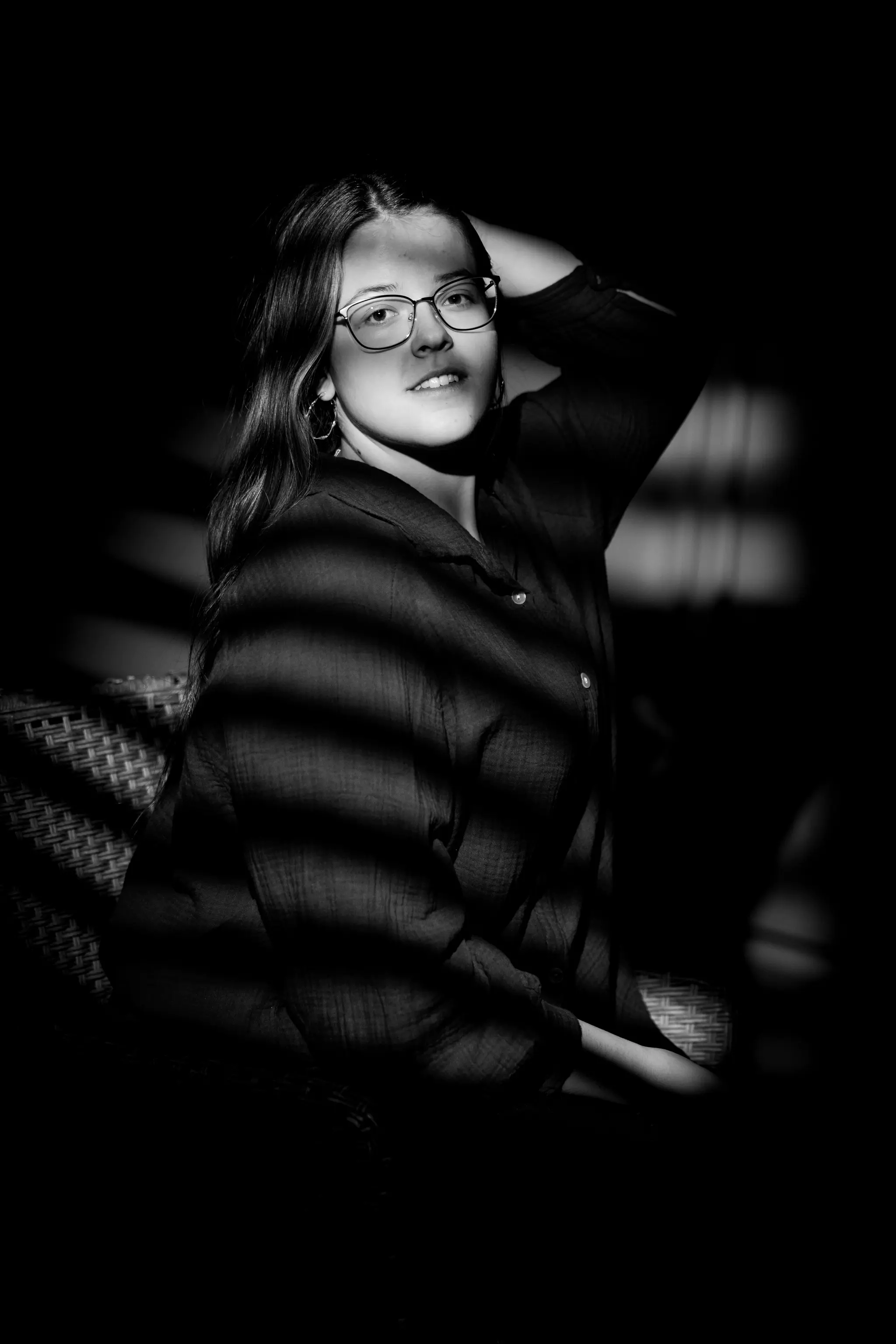 Ein nachdenkliches Schwarz-Weiß-Porträt von Lisa Matschl, die mit Brille und langen Haaren im dramatischen Licht von Schattenstreifen sitzt