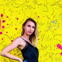 Eine junge Frau mit blonden Haaren und blauen Augen posiert vor einer leuchtend gelben, strukturierten Wand mit pinken Farbspritzern lens: 56mm F1.4 DC DN | Contemporary 018