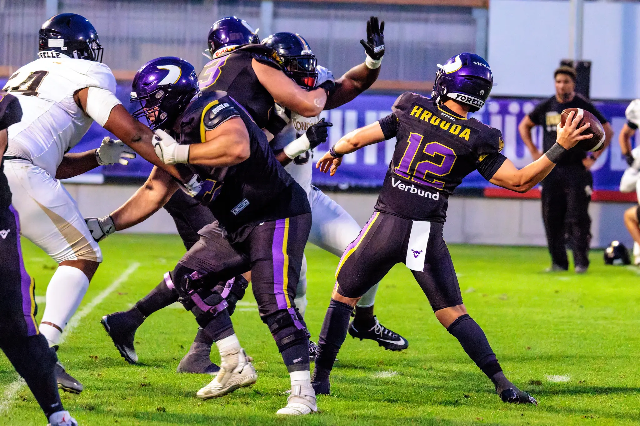 Nico Hrouda (12) setzt unter Druck zum knappen Pass an, während die Offensive Line der Vienna Vikings die Londoner Pass-Rusher blockt – enger Spielzug im vierten Viertel.
