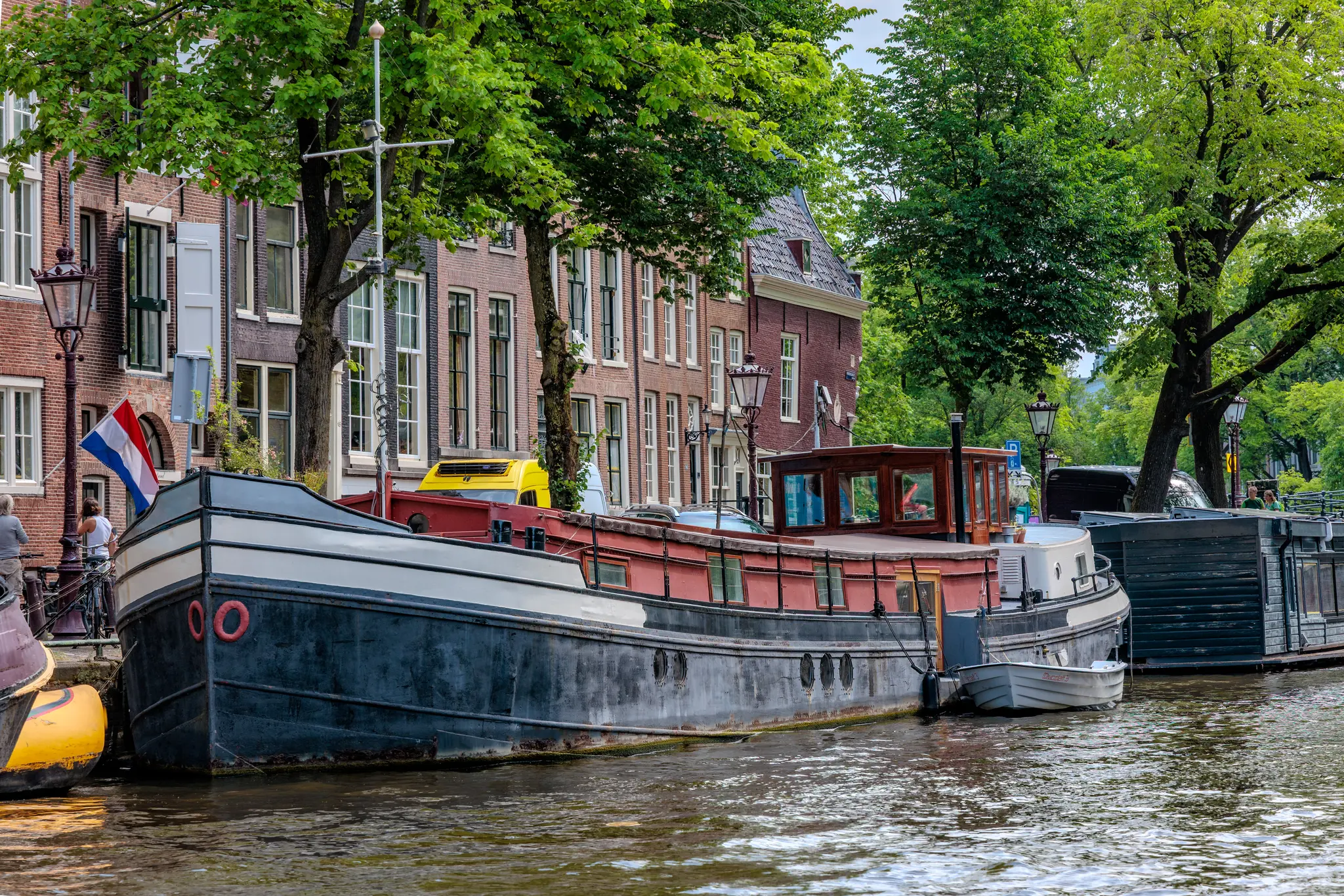 Ein malerisches Amsterdamer Grachtenmotiv mit einem Hausboot vor klassischen Ziegelhäusern und sattgrünen Bäumen 