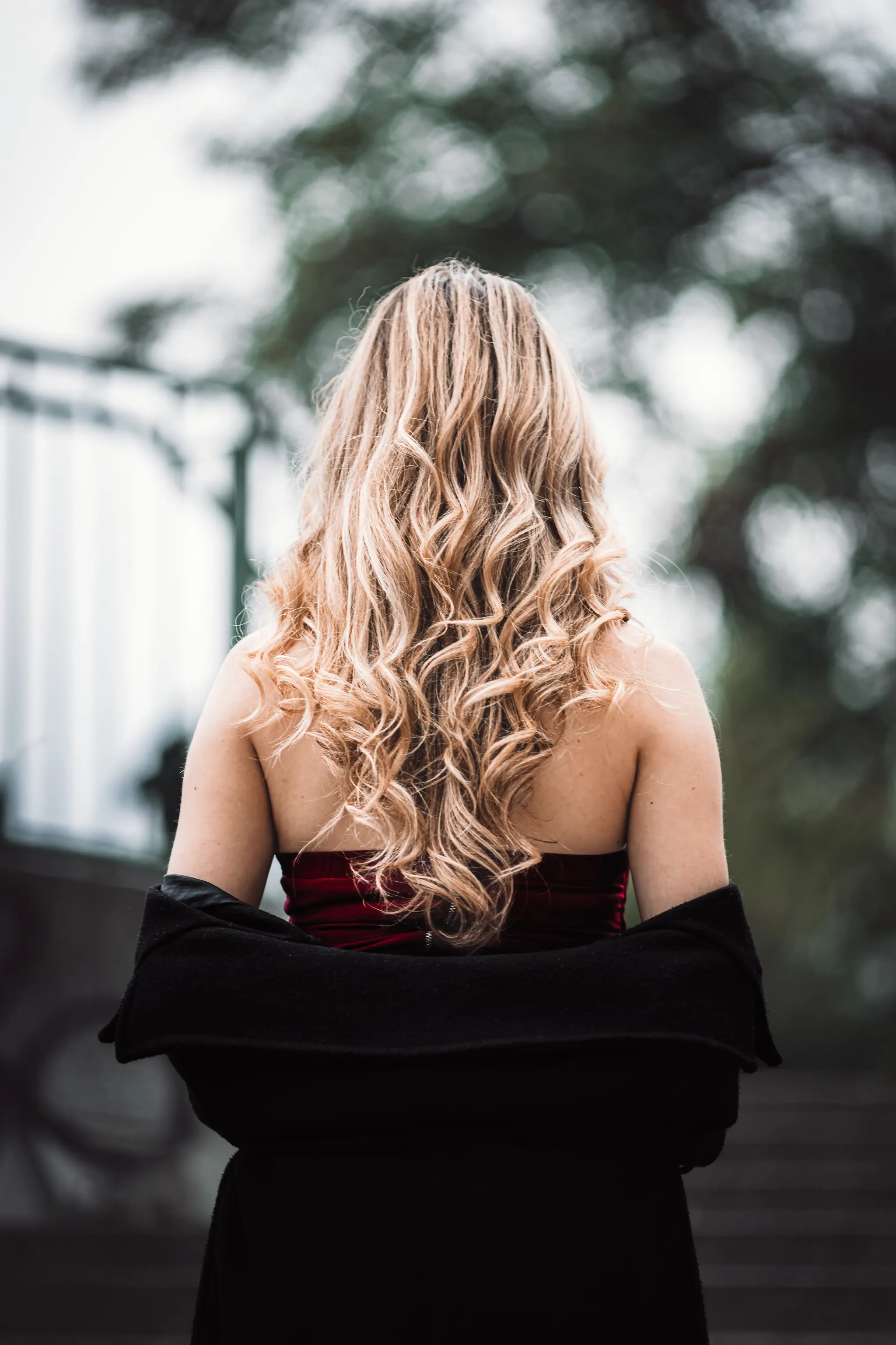Rückansicht von Model Valentina mit langen, blonden Locken, die während ihres Porträt-Shootings im Wiener Stadtpark ein rotes Kleid und einen schwarzen Mantel trägt