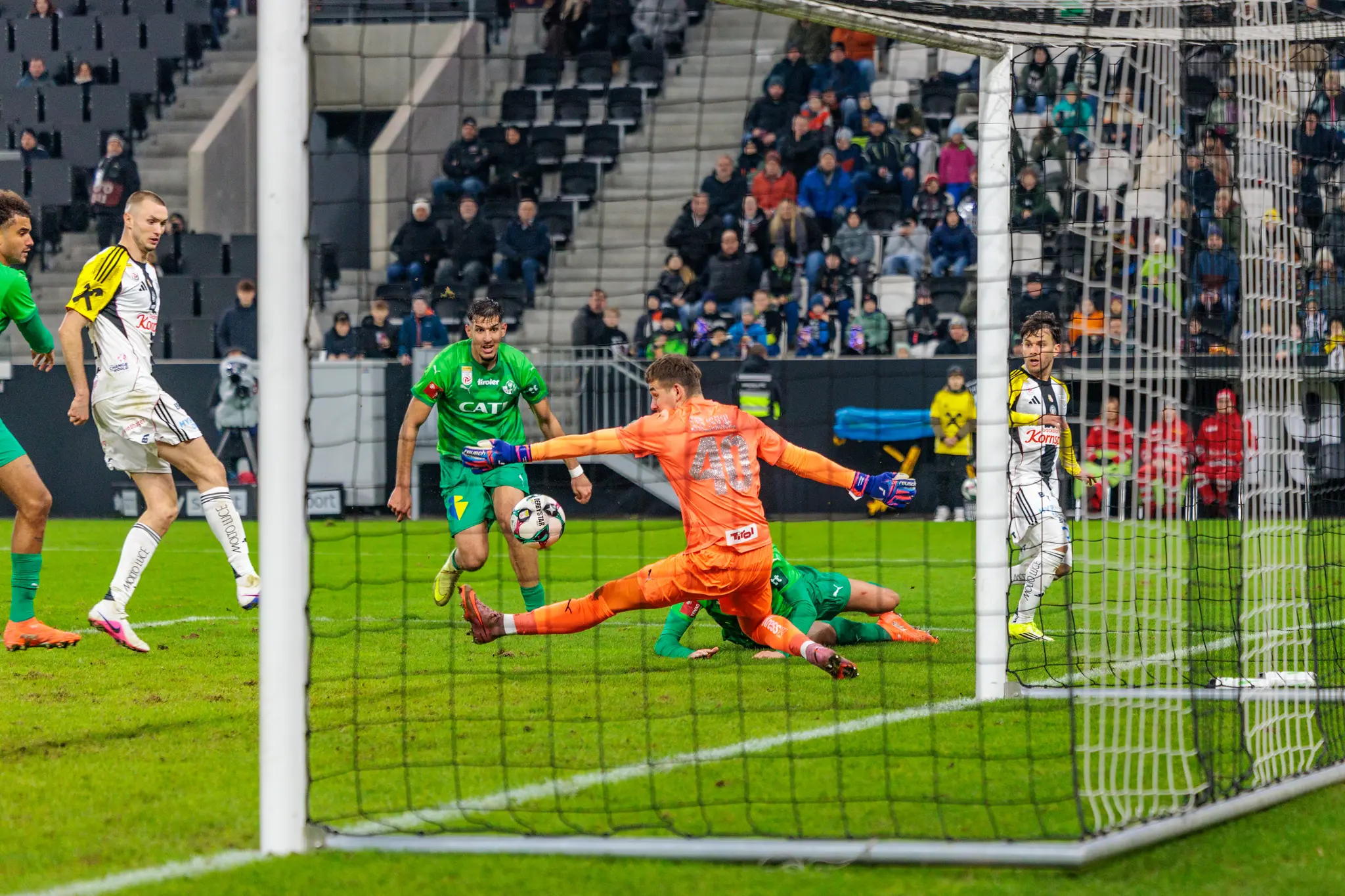 Ein orangefarbener Fußballtorhüter pariert mit ausgestreckten Armen einen Schuss, während ein Spieler in grünem Trikot nahe des Balles im Strafraum agiert