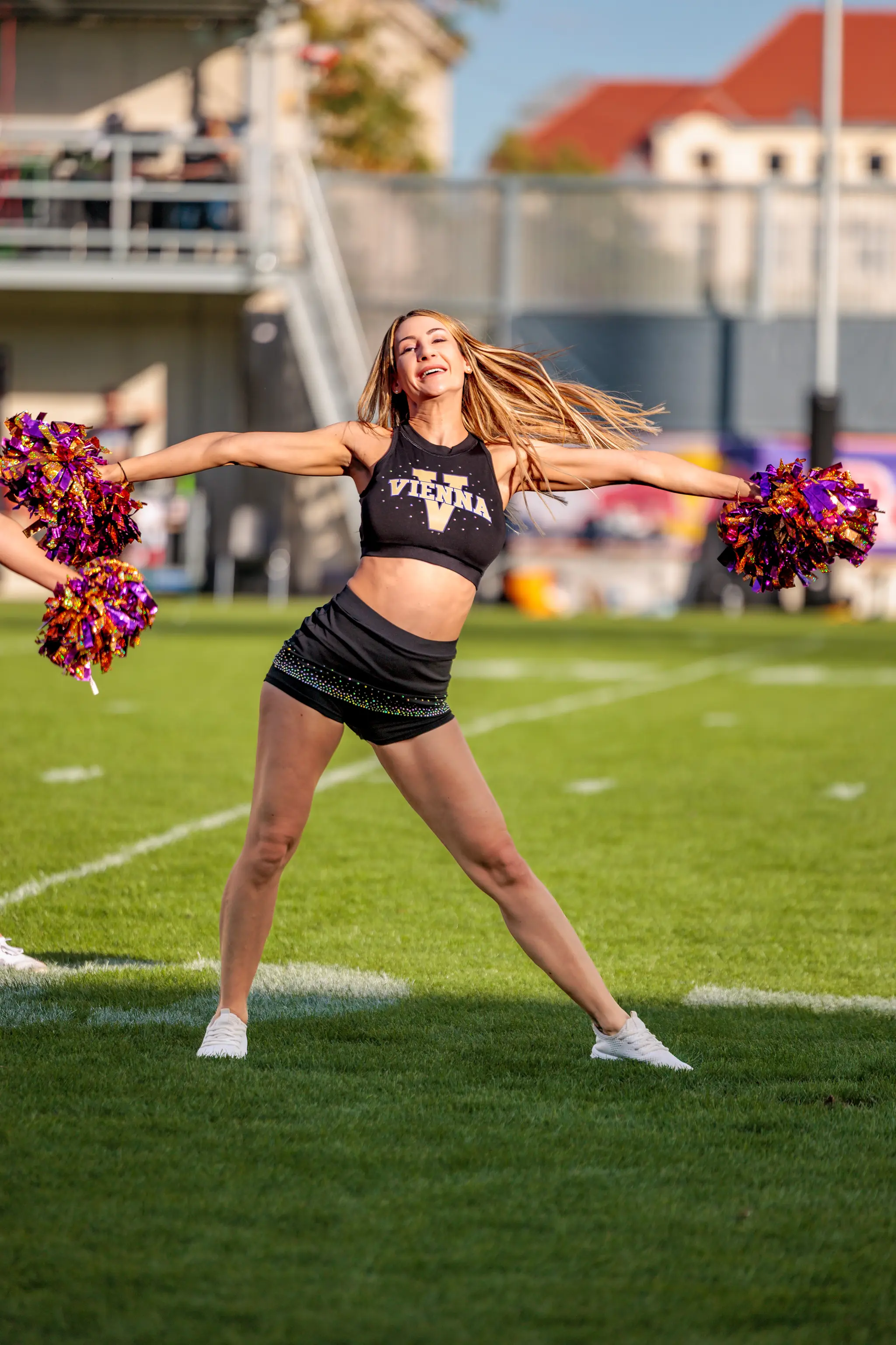 Vor dem Spiel zeigt eine Tänzerin des Vienna Vikings Cheer Squads mit ausgebreiteten Armen und lila-goldenen Pompons eine lebhafte Pose auf dem Rasen im Wiener Sport-Club-Stadion.