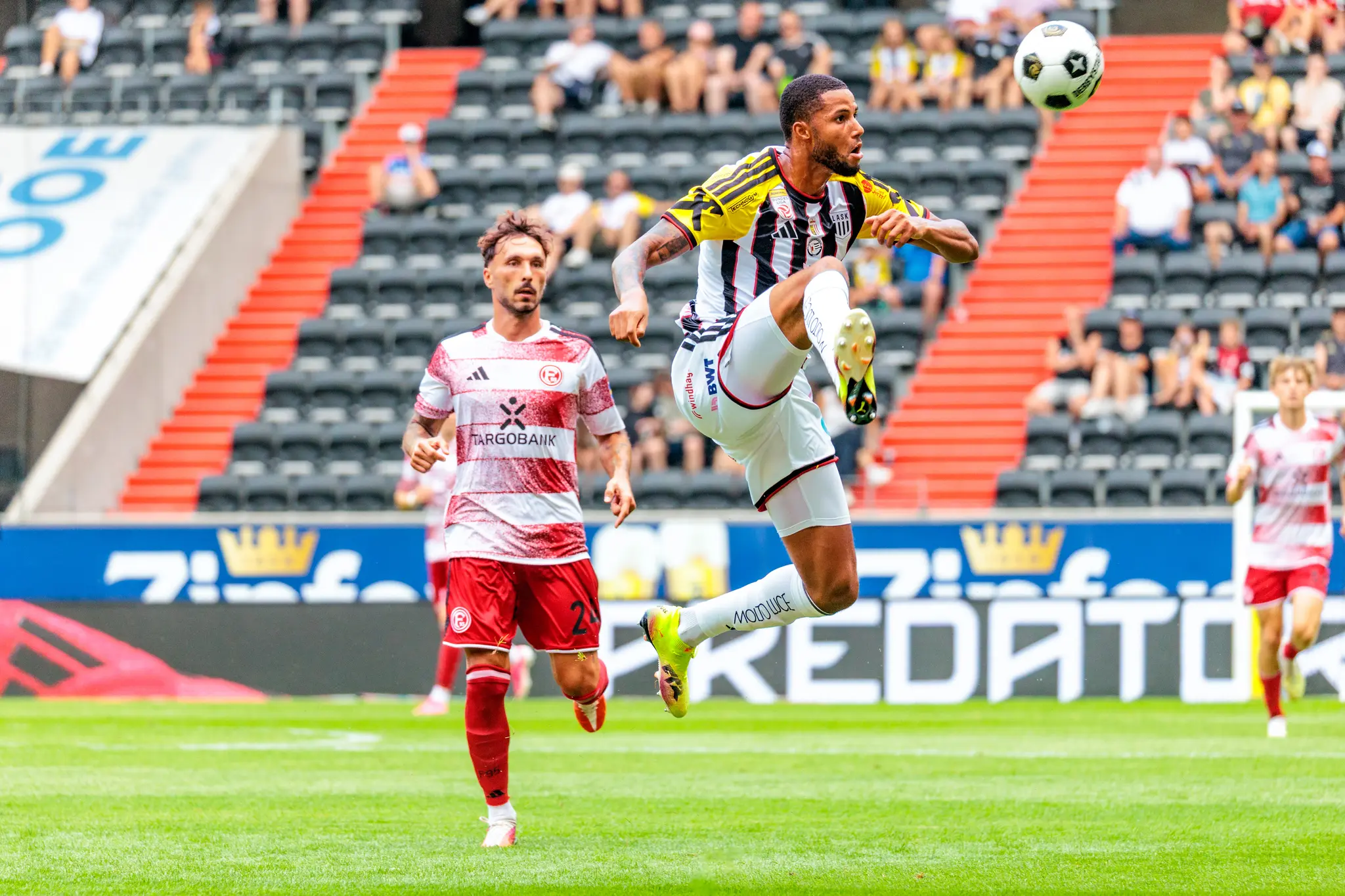 LASK-Spieler nimmt den Ball artistisch mit dem Fuß im Sprung an, ein Düsseldorfer Spieler steht im Vordergrund.