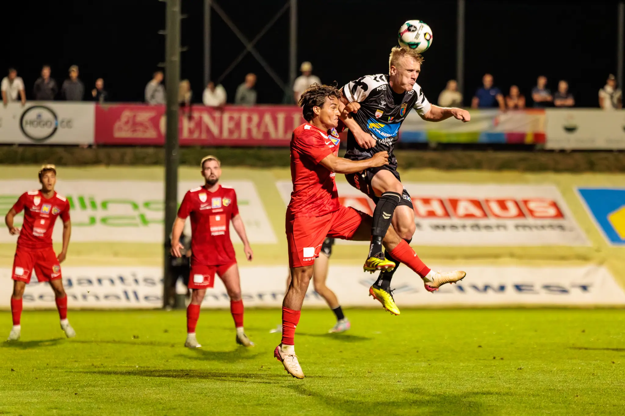 Ein intensiver Kopfball-Zweikampf zwischen zwei Fußballspielern in der Luft während eines abendlichen Matches, bei dem der Spieler von Herta Wels den Ball köpft