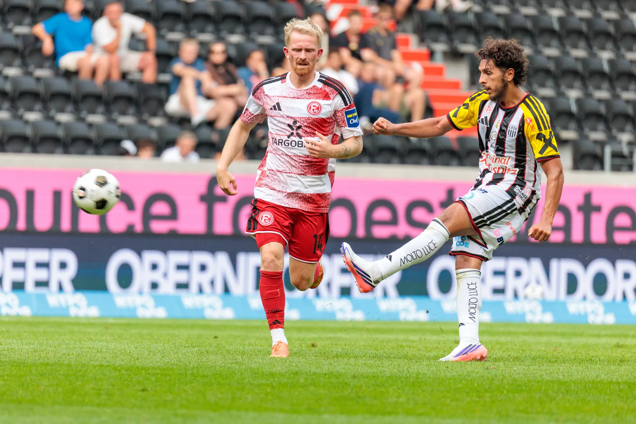 LASK-Spieler streckt das rechte Bein zum Ball, während ein Düsseldorfer Verteidiger herannaht, der Ball fliegt in der Luft zwischen beiden.