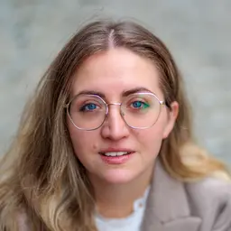 Nahaufnahme einer lächelnden jungen Frau mit blonden Haaren und modischer Brille, deren leuchtend blaue Augen hervorstechen