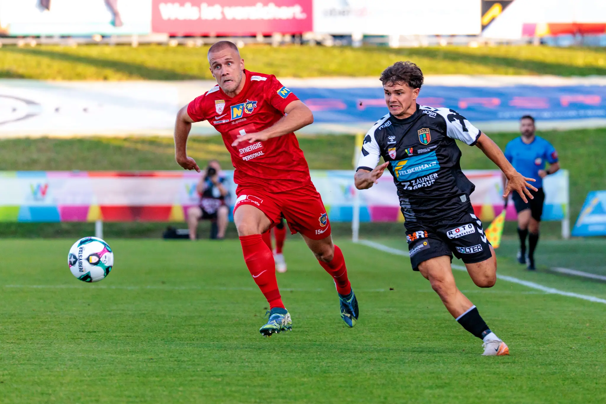 Im ÖFB Cup kämpfen ein Spieler des SKN St. Pölten in rotem Trikot und ein Kontrahent von Herta Wels in schwarz-weißem Dress energisch um den Ball