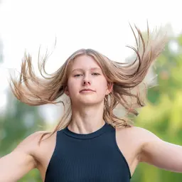Porträt einer jungen Frau mit dynamisch wehendem blondem Haar und dunklem Oberteil vor einem hellen, unscharfen Hintergrund lens: 56mm F1.4 DC DN | Contemporary 018