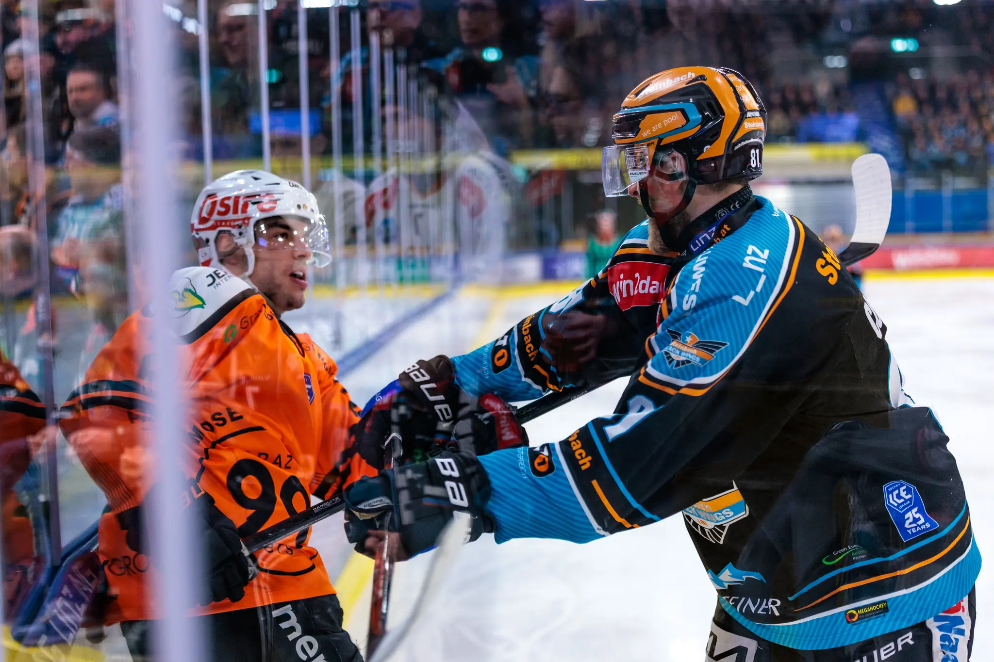 Zwei Eishockeyspieler auf dem Eis, ein Spieler der Graz 99ers in orange und ein Spieler der Black Wings Linz in schwarz-blau, die sich während eines Spiels duellieren 