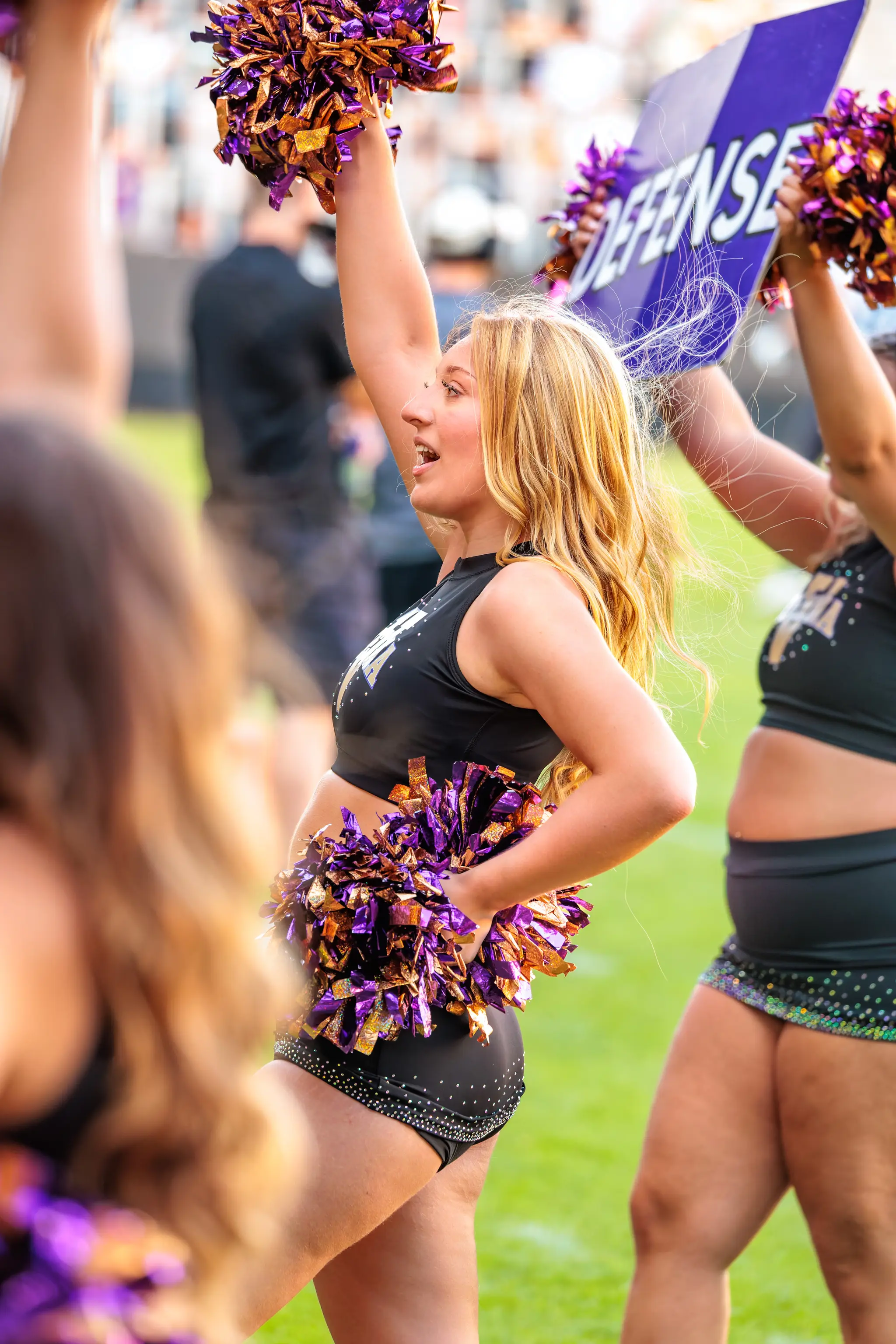 Cheerleaderin des Spirit Squad tanzt mit violett-goldenen Pompons, einer ruht an der Hüfte.
