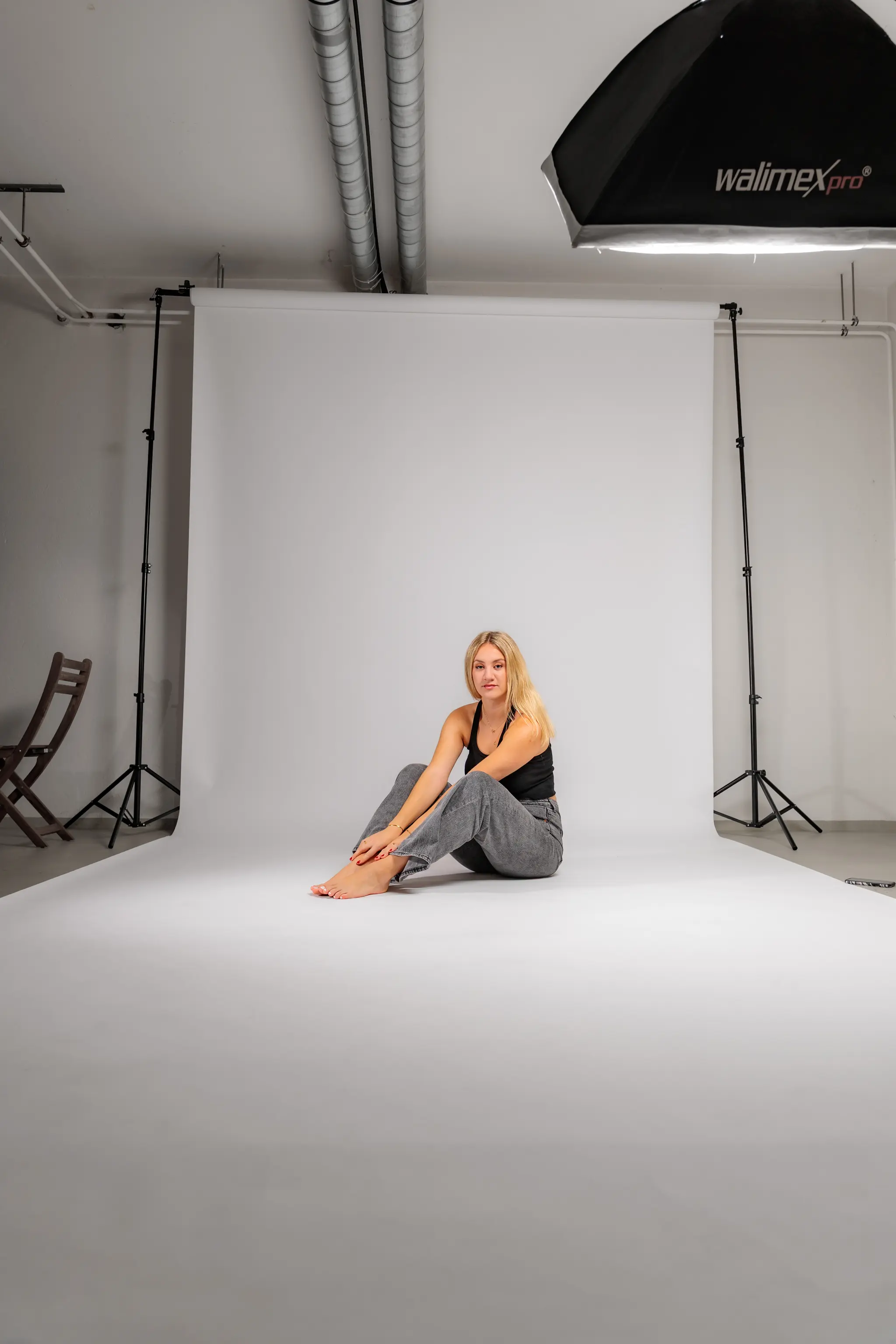 Ein Blick hinter die Kulissen eines Studio-Fotoshootings zeigt Model Anna, die barfuß auf einer weißen Hohlkehle sitzt, umgeben von Stativen und Beleuchtungstechnik