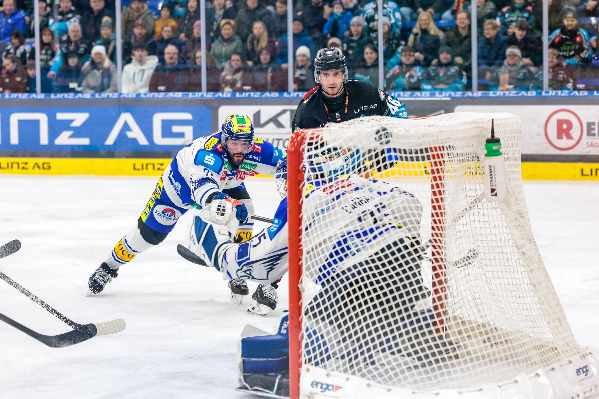 Ein Eishockey-Spieler der Black Wings Linz im blau-weißen Trikot versucht, den Puck am VSV-Torwart in weißer Ausrüstung vorbei ins Tor zu schießen