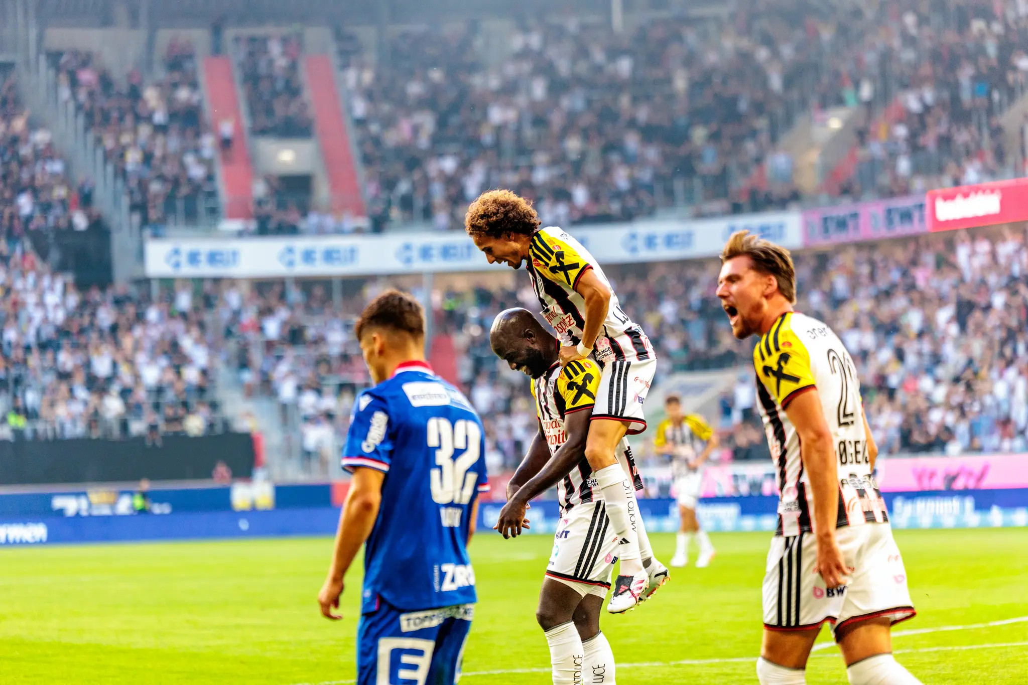 LASK-Spieler feiern ein Tor gemeinsam auf dem Spielfeld, während ein GAK-Spieler enttäuscht davonläuft