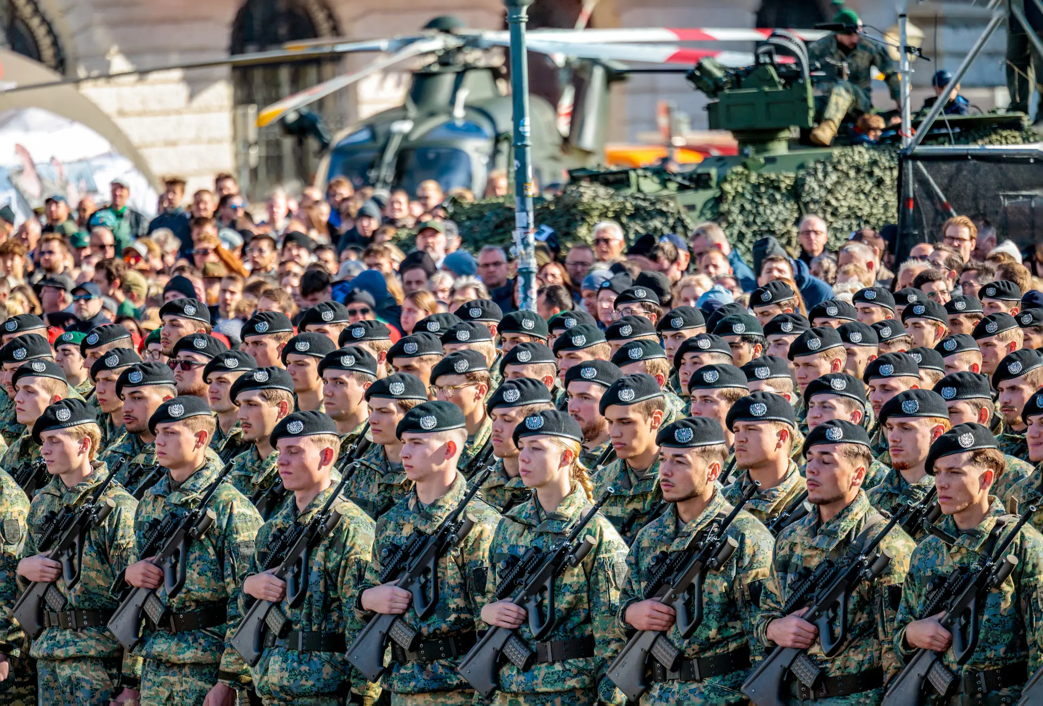 Eine Formation von Rekruten steht mit Sturmgewehren vor einem Hubschrauber und einem Panzer, dahinter verfolgt das Publikum die Angelobung.