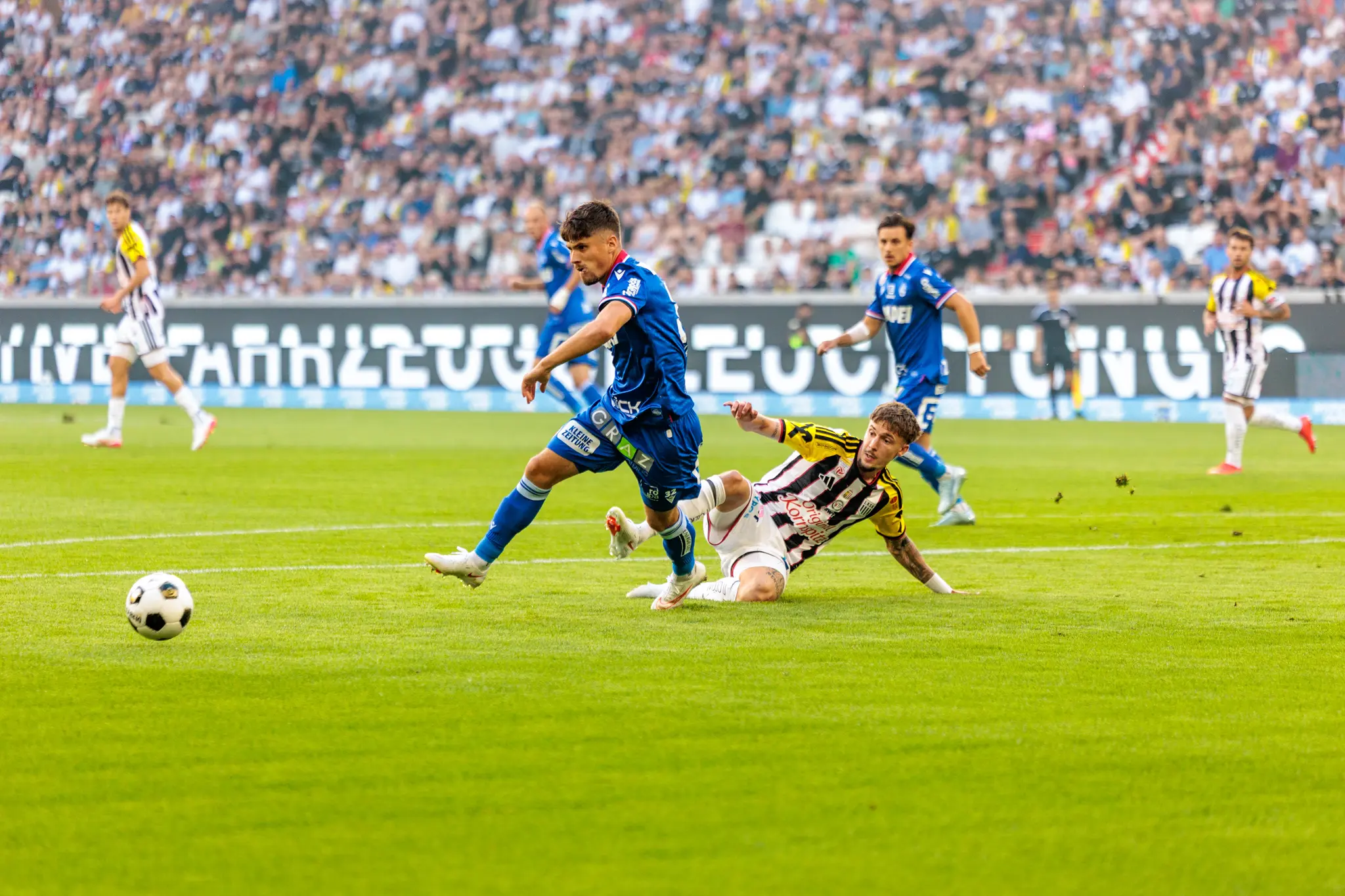 LASK-Spieler rutscht am Boden, während ein GAK-Spieler im blauen Trikot herannaht und die Spielsituation im Mittelfeld ausgetragen wird