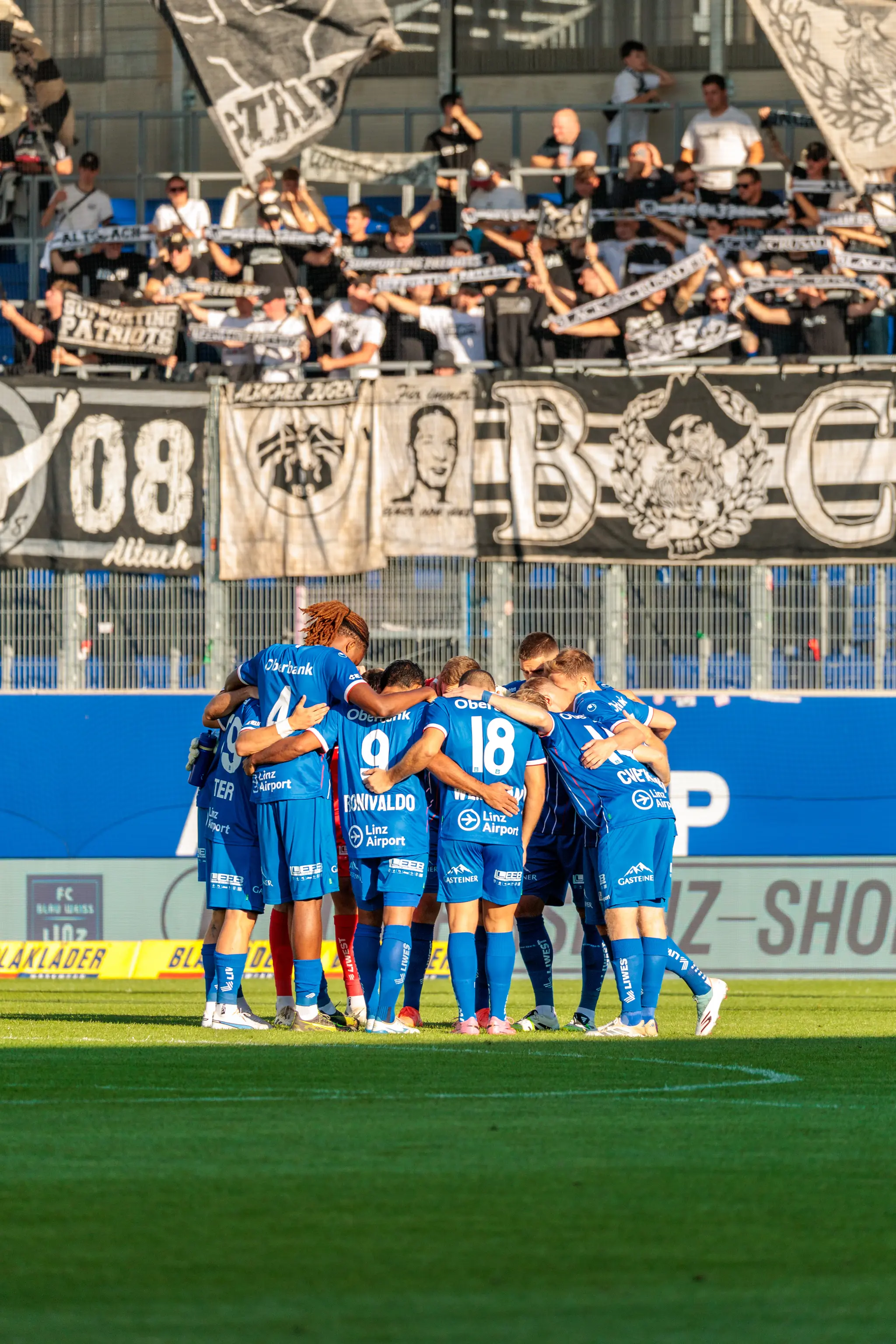 Die Fußballmannschaft des FC Blau-Weiß Linz bildet einen engen Kreis zur Teambesprechung vor der Fankurve mit den gegnerischen Zuschauern