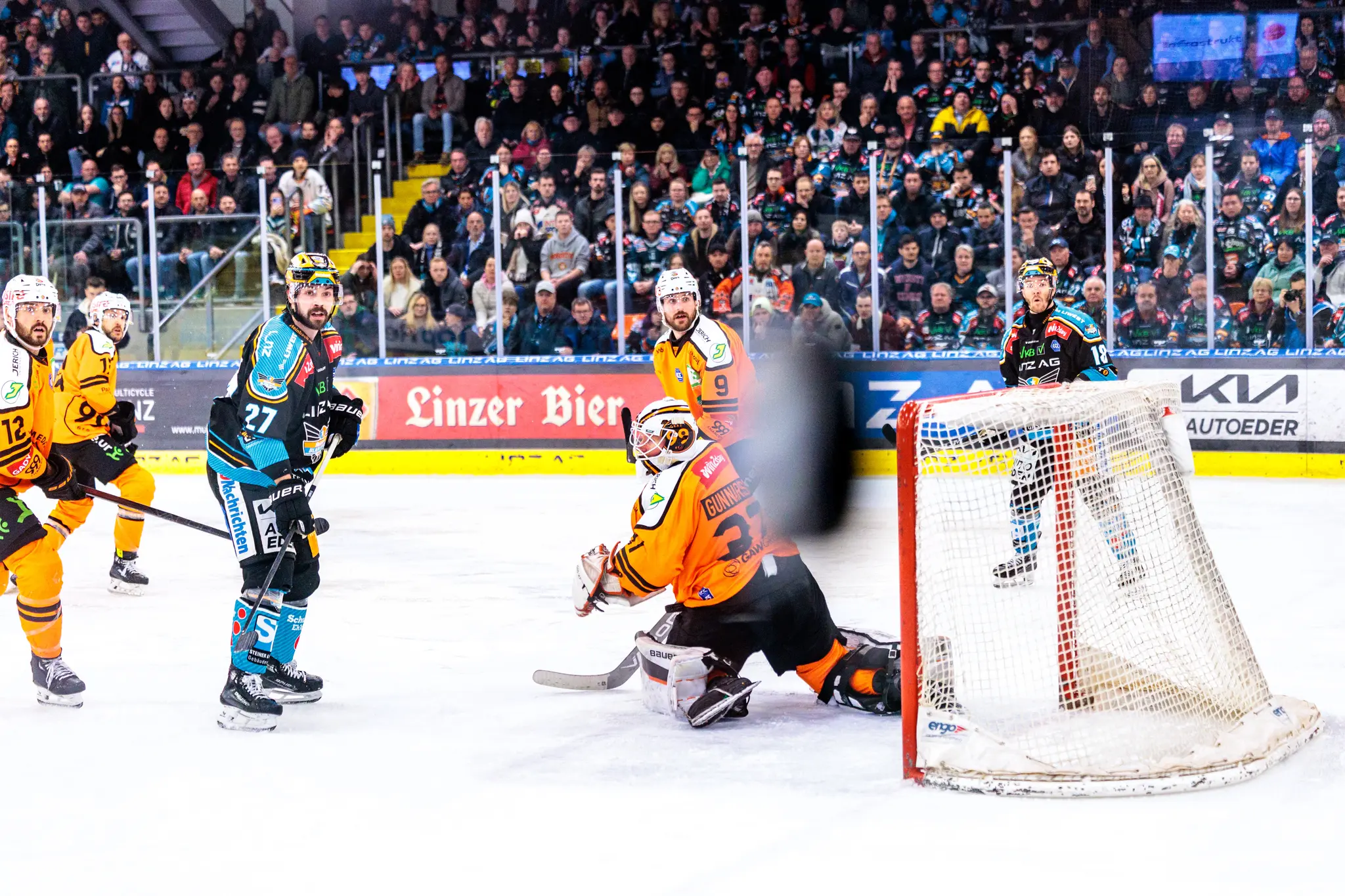 Ein Eishockeytorwart der Grazer 99ers blickt während eines Spielzugs vor seinem Tor in Richtung Aktion, umgeben von Spielern ibeider Mannschaften- ein unscharfer Eishockeypuck im Vordergrund an der Scheibe 
