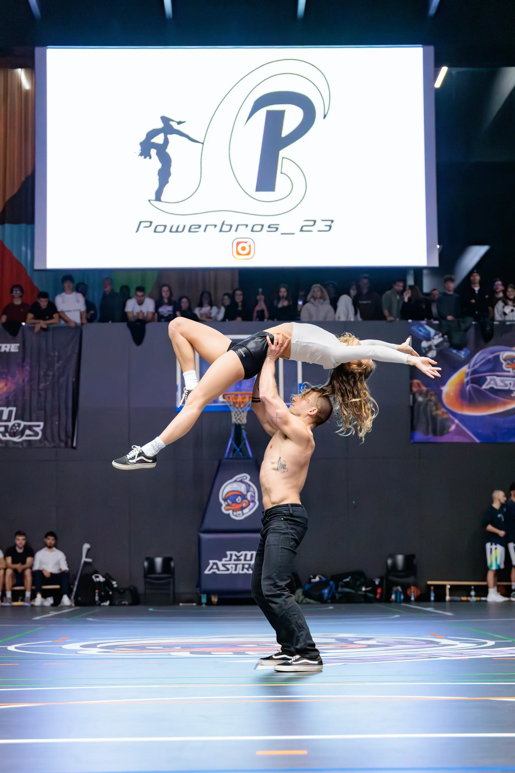 Zwei Athleten führen auf einem Sportplatz eine beeindruckende akrobatische Hebefigur vor, während im Hintergrund ein großer Bildschirm das Logo „Powerbros_23“ und Zuschauer zeigt 
