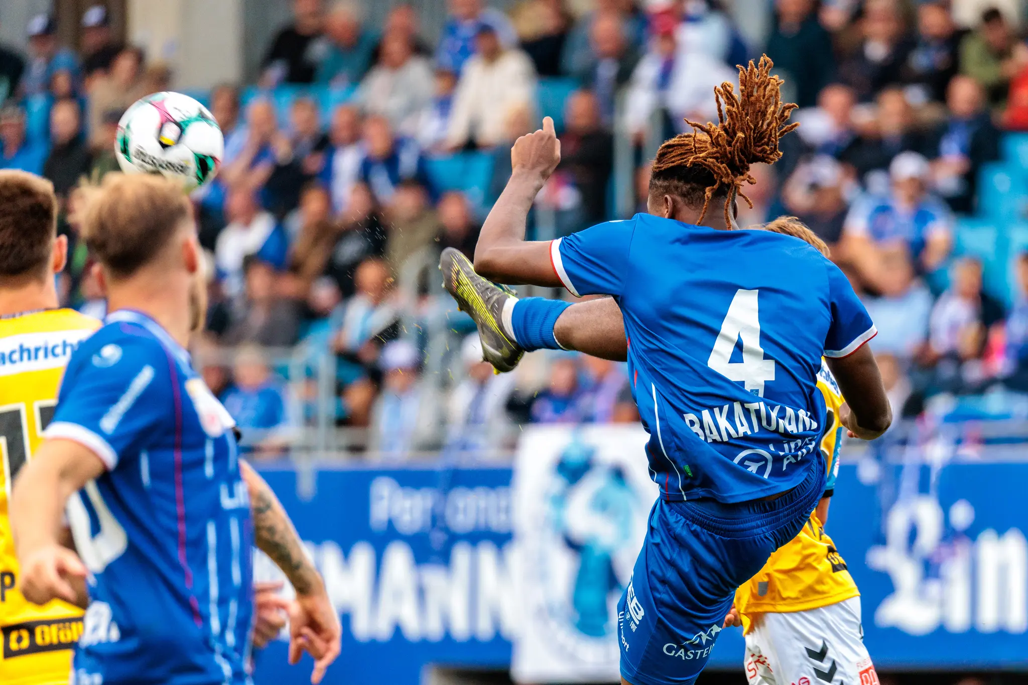 Ein Verteidiger von Blau-Weiß Linz mit der Rückennummer 4 und Dreadlocks klärt den Fußball mit einem kräftigen Schuss im Spiel gegen SV Ried vor der unscharfen Kulisse der Zuschauer